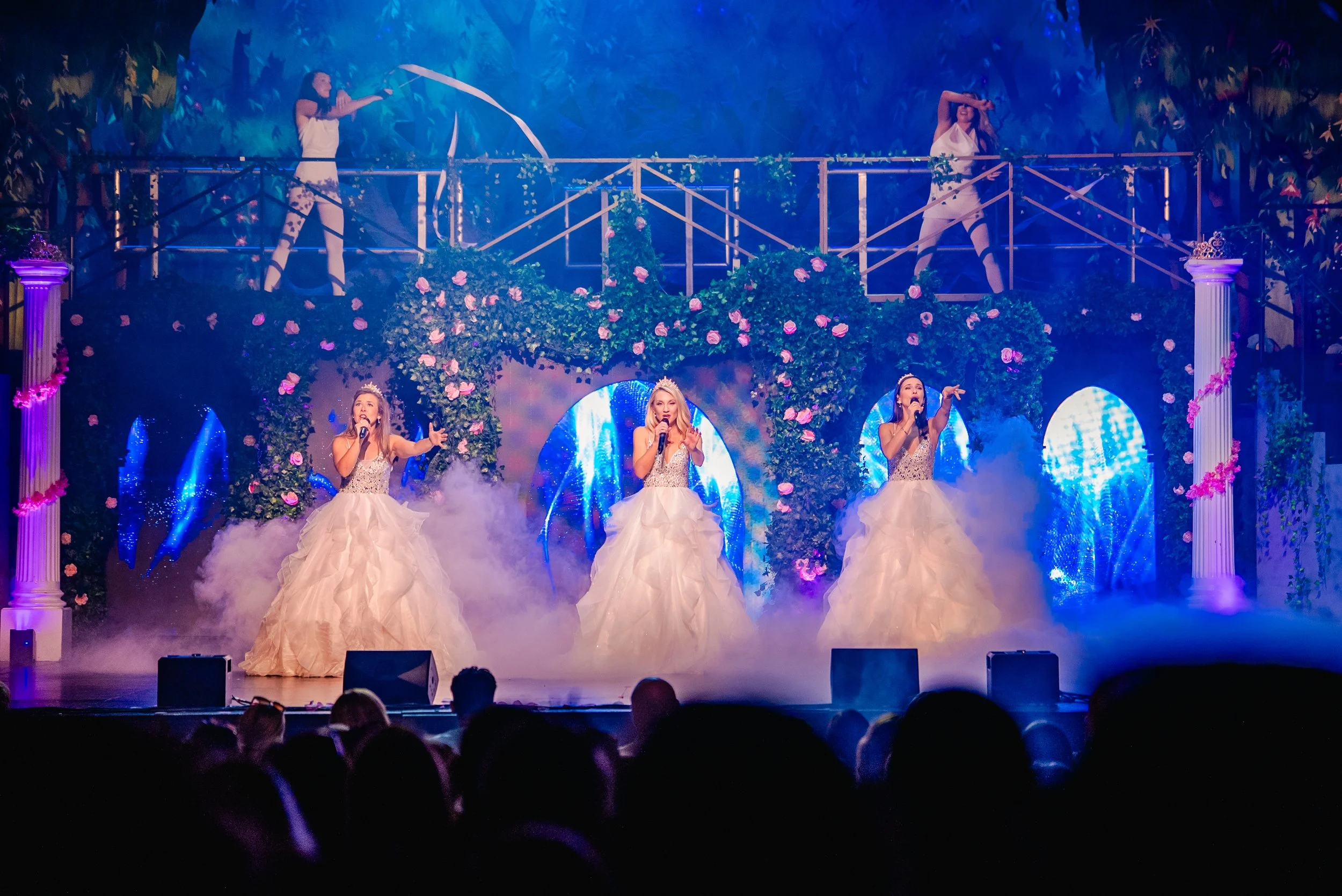 Three female singers in white ball gown dresses perform on stage with a colorful, fantasy-themed backdrop, including flowers, trees, and columns, with an audience watching.