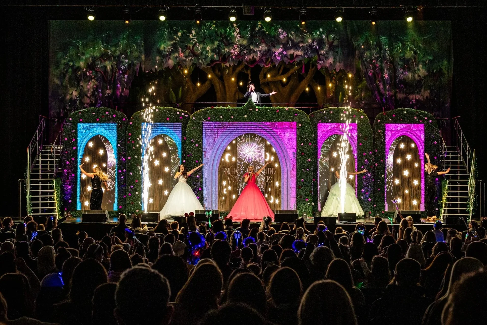 A stage performance featuring a male singer on an elevated platform and four female singers on the main stage, with colorful backdrops, decorative arches, and sparking streamers, attended by a large audience.