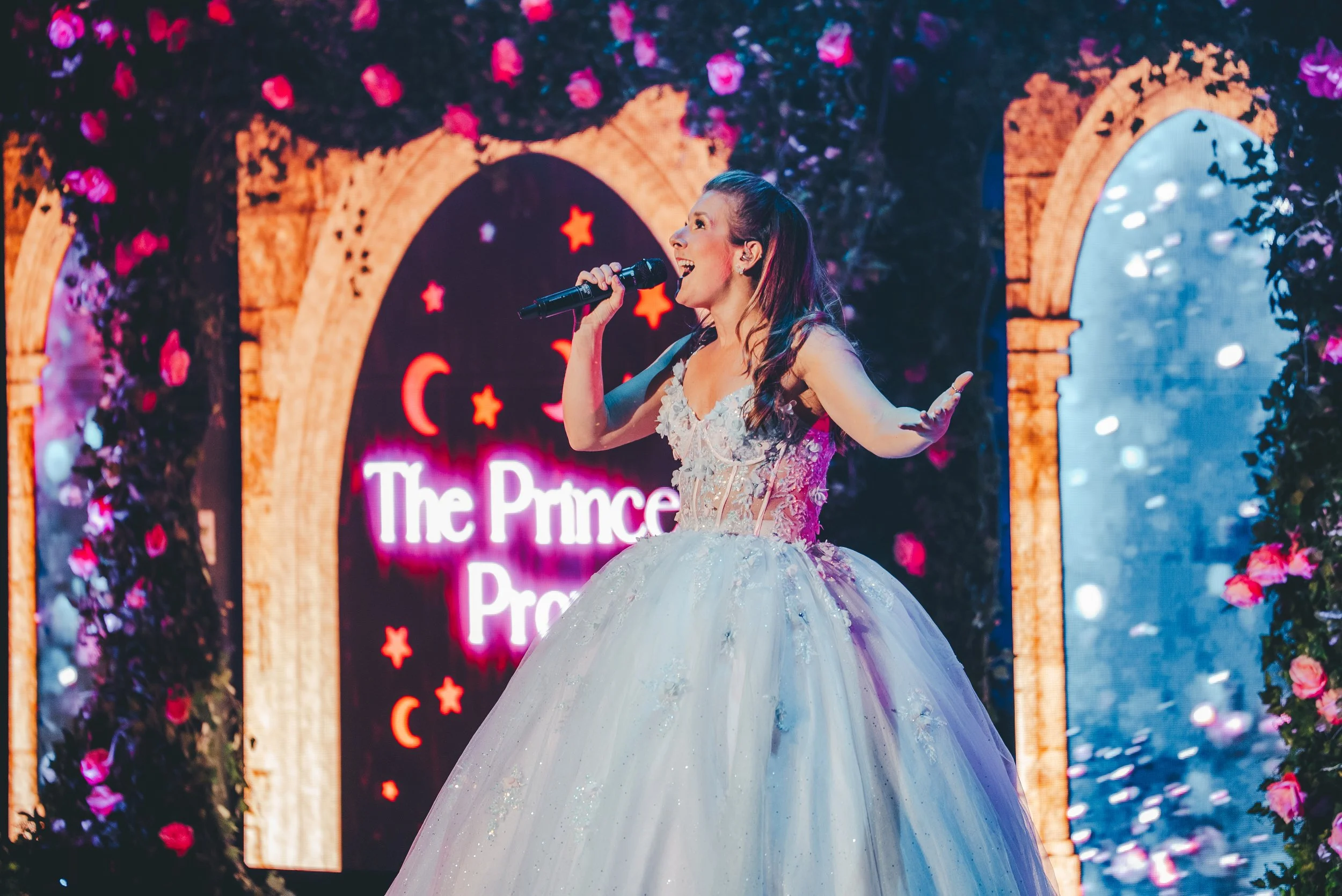 A woman in a strapless, white, flower-adorned ball gown singing into a handheld microphone on a decorated stage with floral arch and screens displaying stars and crescent moons in the background.