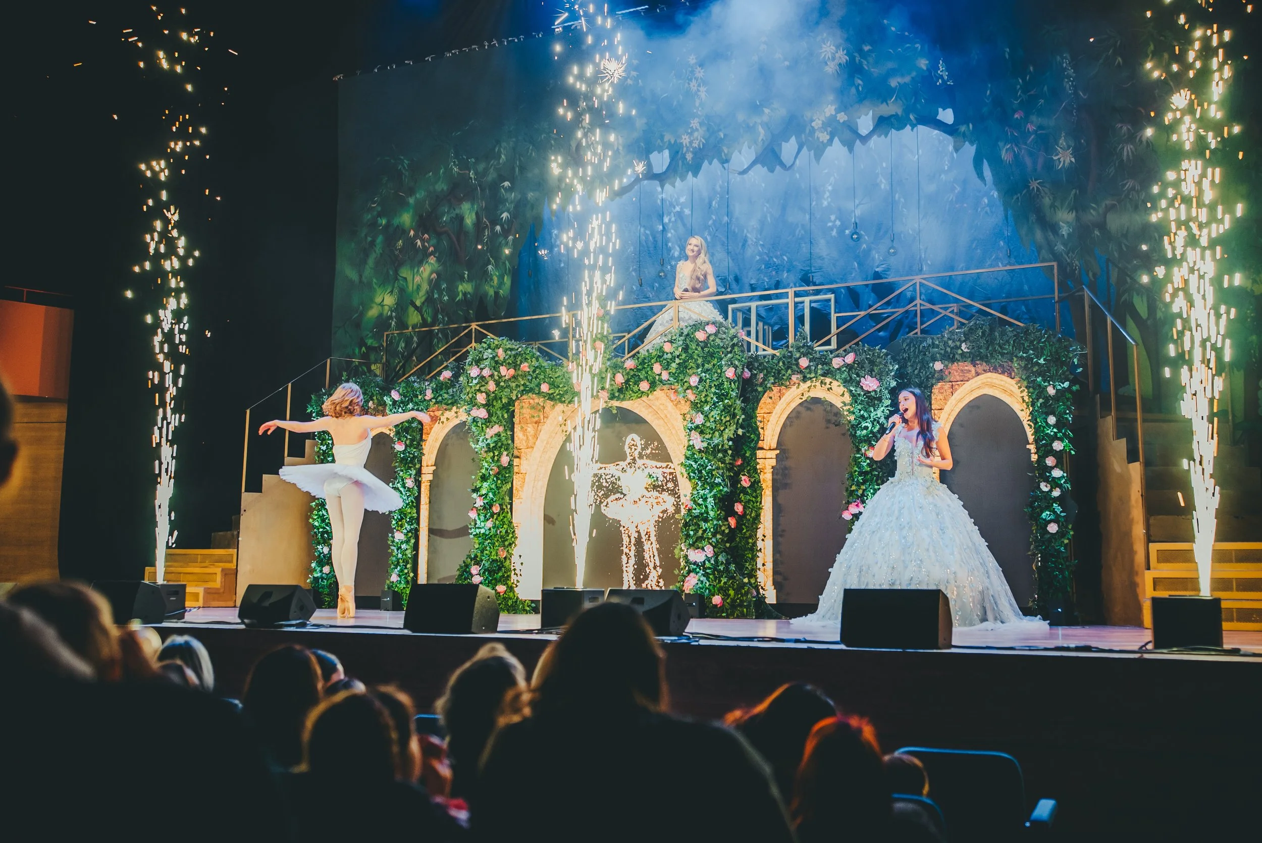A stage performance with a woman in a white dress with a full skirt and floral embellishments, singing into a microphone. Behind her, a backdrop depicts a lush, green forest scene. There are two other women in ballerina costumes and fireworks or pyrotechnics are erupting on the left and right sides of the stage. The audience's silhouettes can be seen at the bottom of the image.