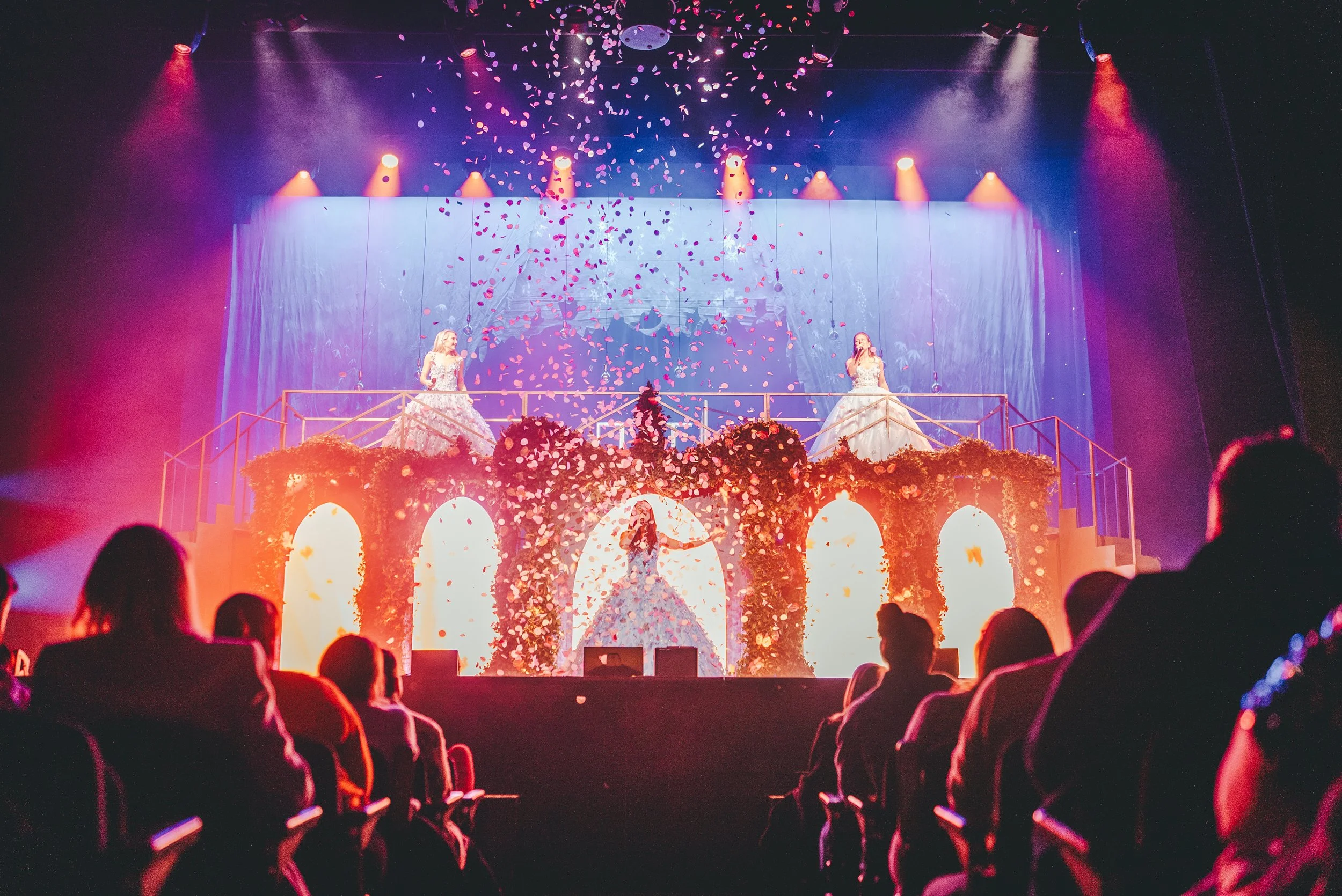 Dance performance on stage with two women in elaborate dresses, surrounded by falling flower petals, with an audience watching.