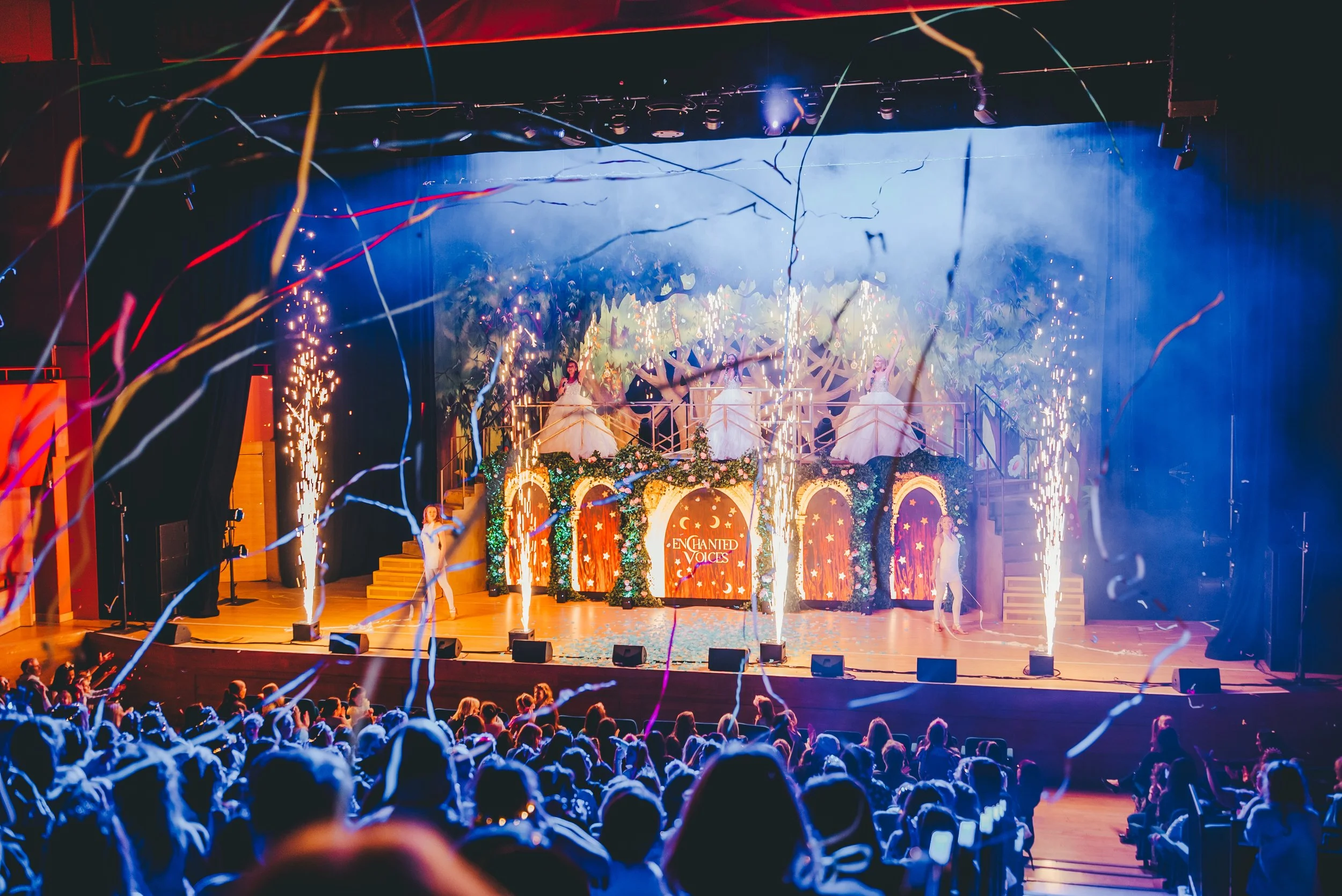 The stage shows a theatrical performance with three women in white dresses, surrounded by colorful lights, fireworks, and streamers. The backdrop features lush greenery and the words "Enchanted Voices" with a moon and star design, indicating a magical or fantasy theme. The audience is visible in the foreground, watching the show.