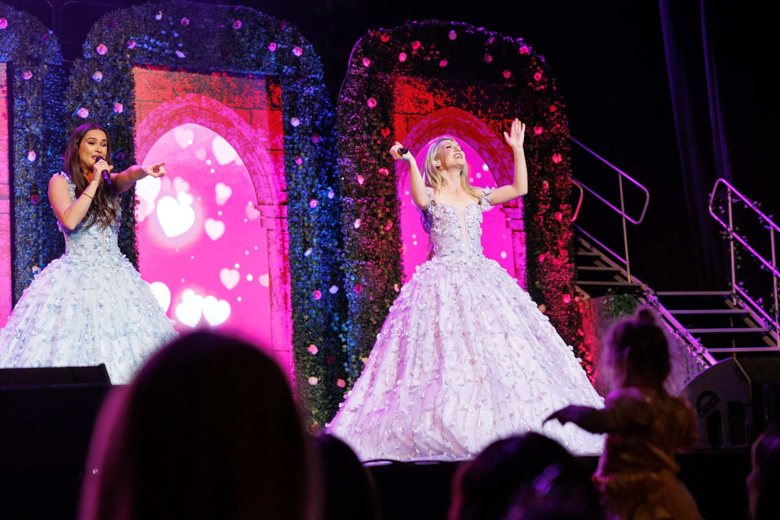 Two women in elaborate white ball gowns performing on stage with pink and purple heart-themed background decorations.