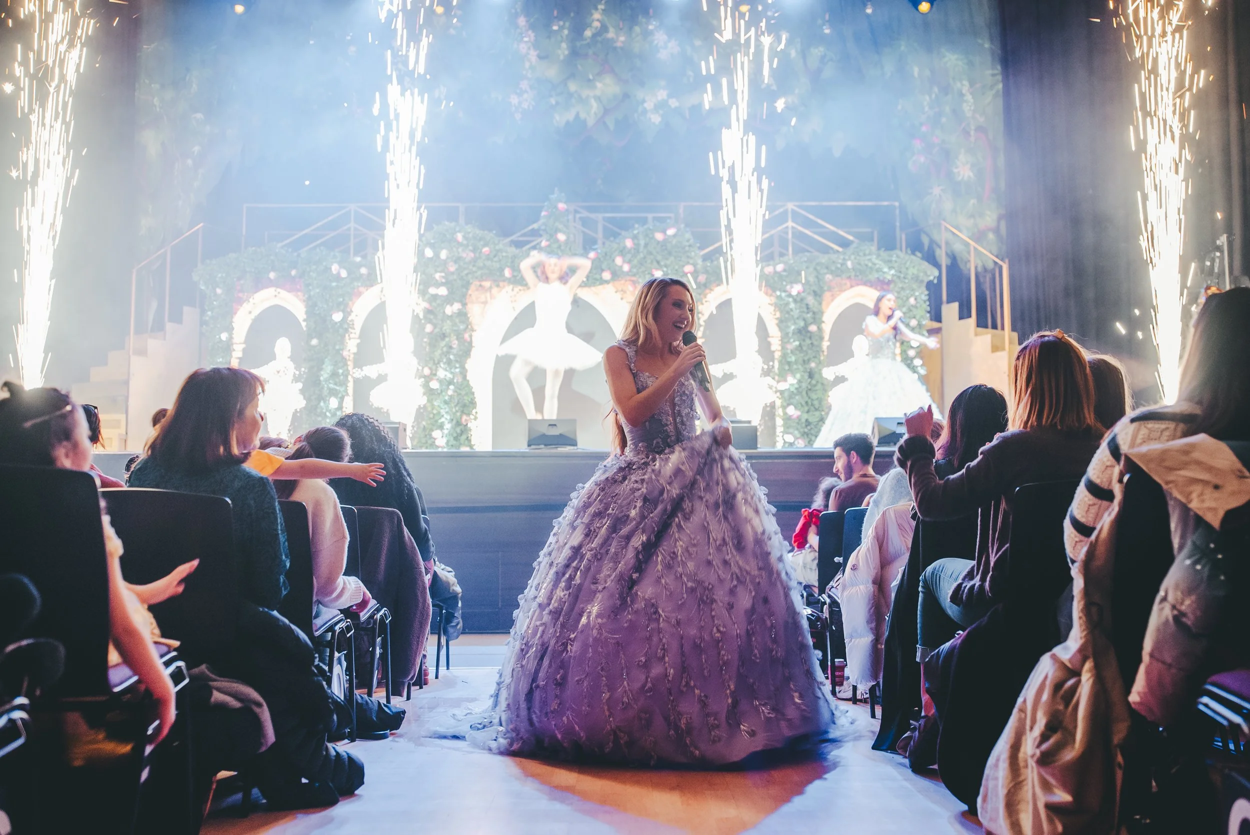 A performer in a purple and white ball gown stands on stage singing into a microphone as sparks fly from the sides of the stage. An audience watches and applauds.
