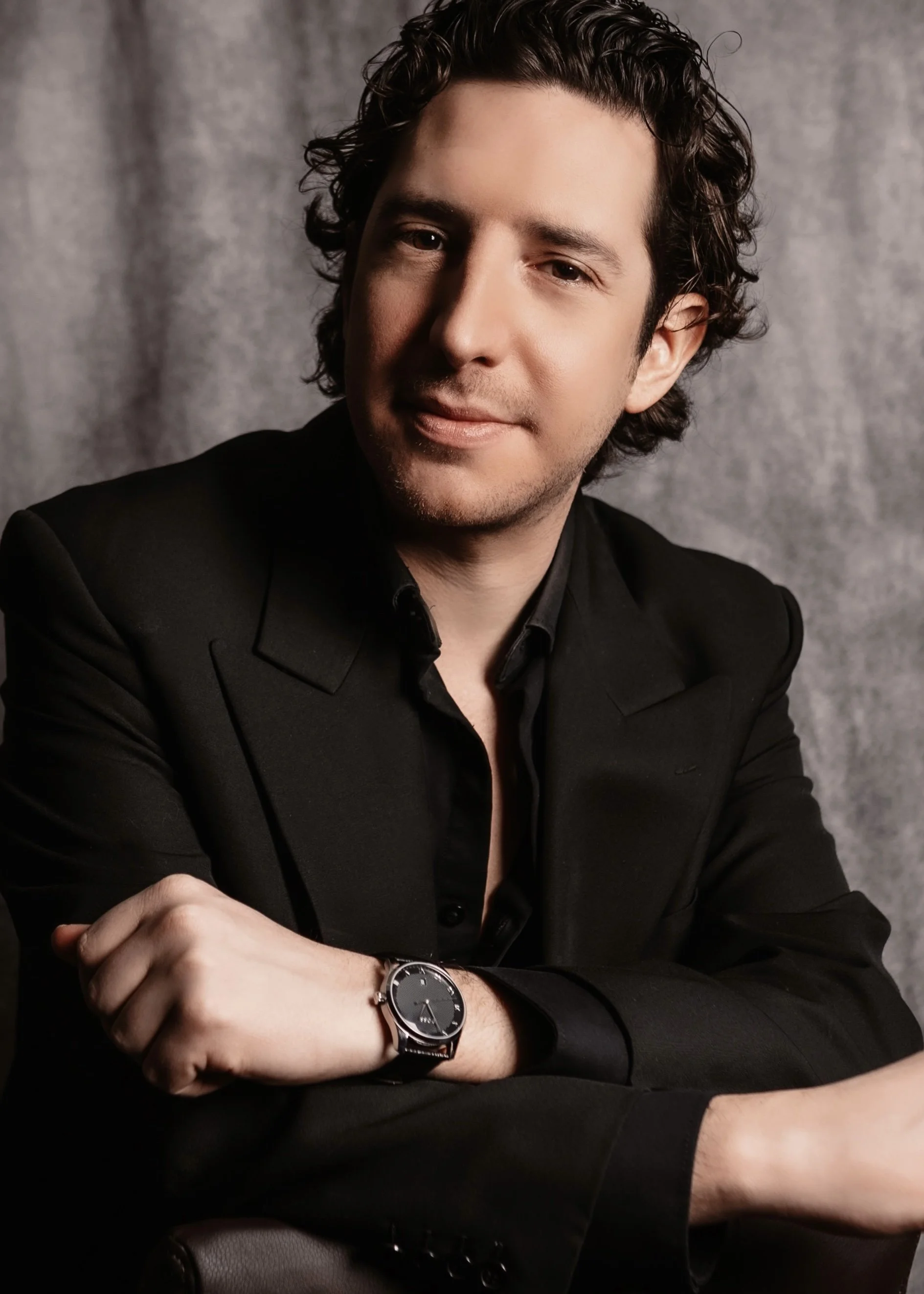 A man with dark curly hair and light skin, wearing a black suit and black shirt, seated with his arms crossed, wearing a black watch, against a gray textured background.