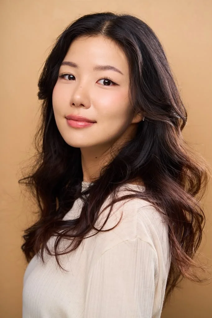 A young woman with long wavy dark hair and a light complexion, looking at the camera with a soft smile, wearing a beige top, against a beige background.