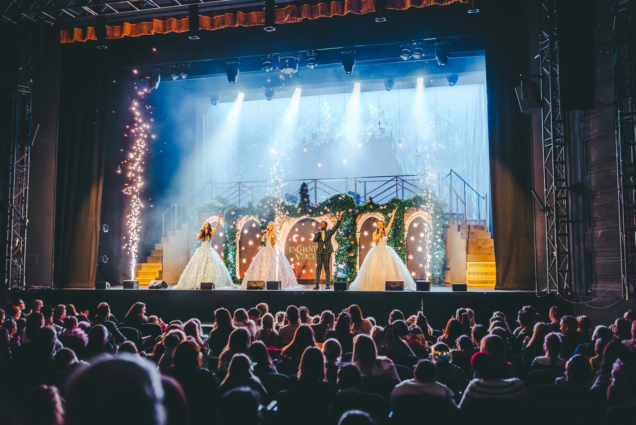 Stage performance with four singers, three women in ball gowns and one man in a tuxedo, under bright blue and white stage lights, with an audience watching in a theater decorated for a festive event.