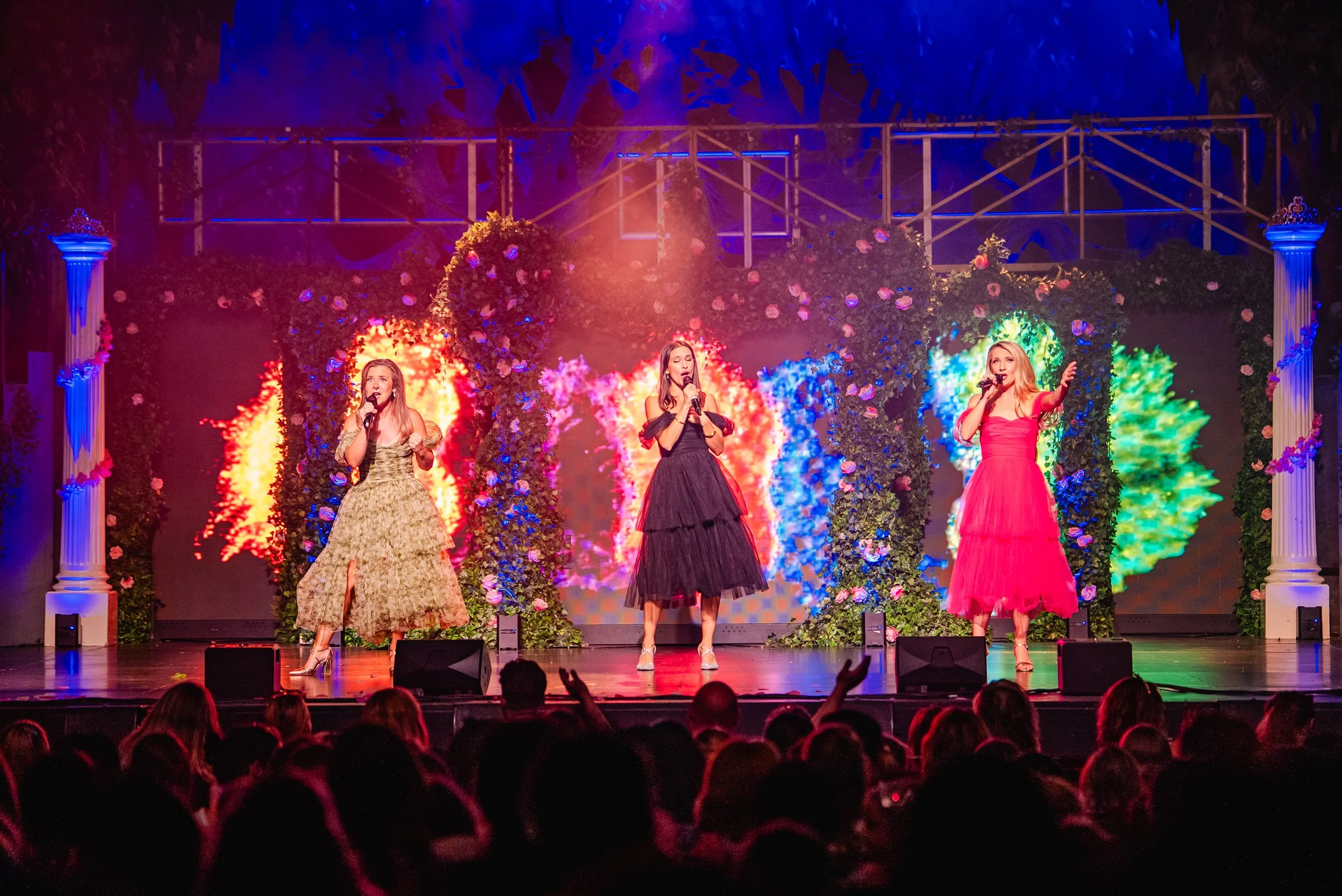 Three women singing on stage during a performance, decorated with colorful floral and abstract backgrounds, illuminated with vibrant stage lights, with an audience visible in the foreground.