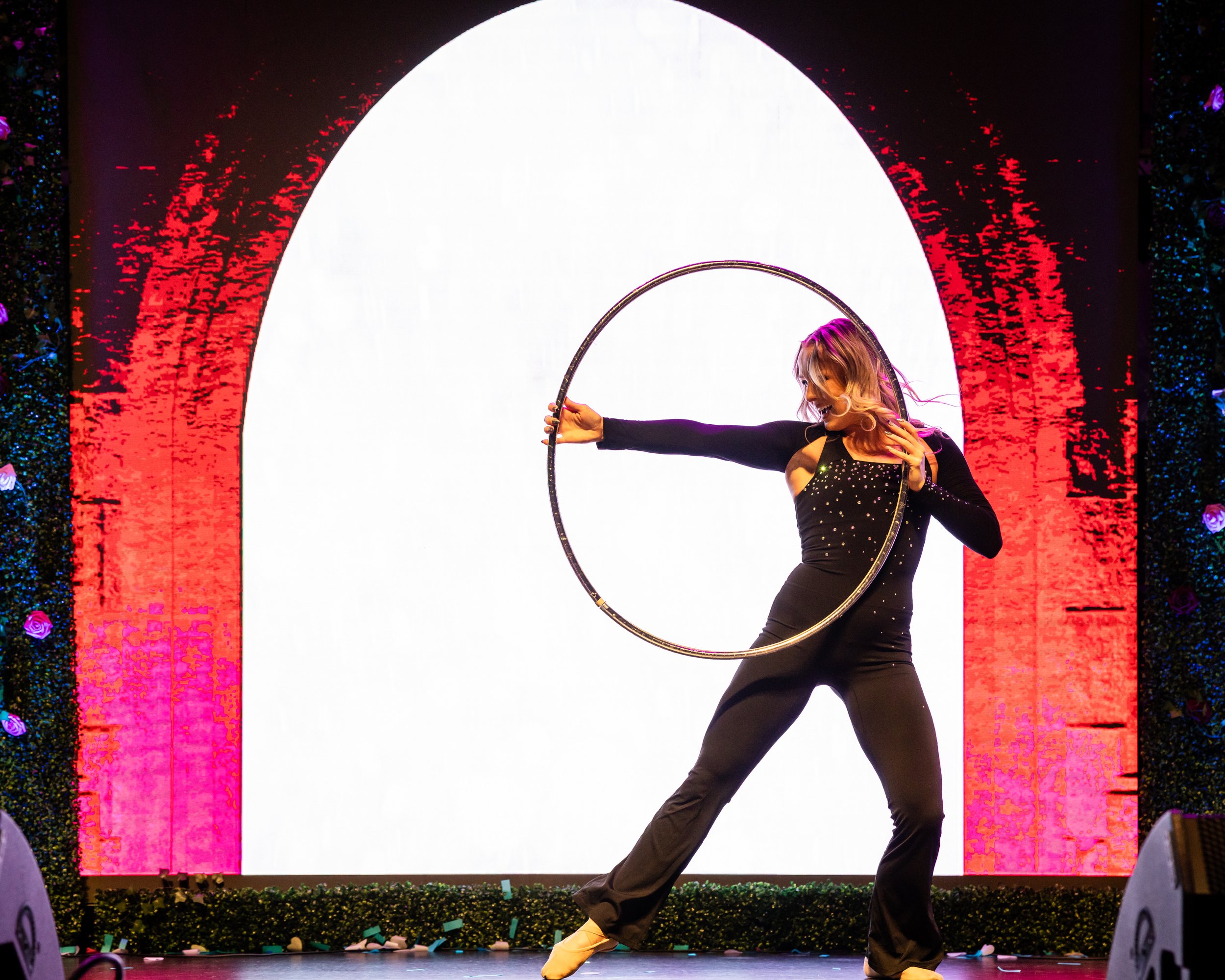 A performer is dancing with a large metal hoop on stage. She is wearing a black dress with sparkling details and yellow shoes. The background features a large white semi-circle with red and dark borders, and decorative elements on the sides.