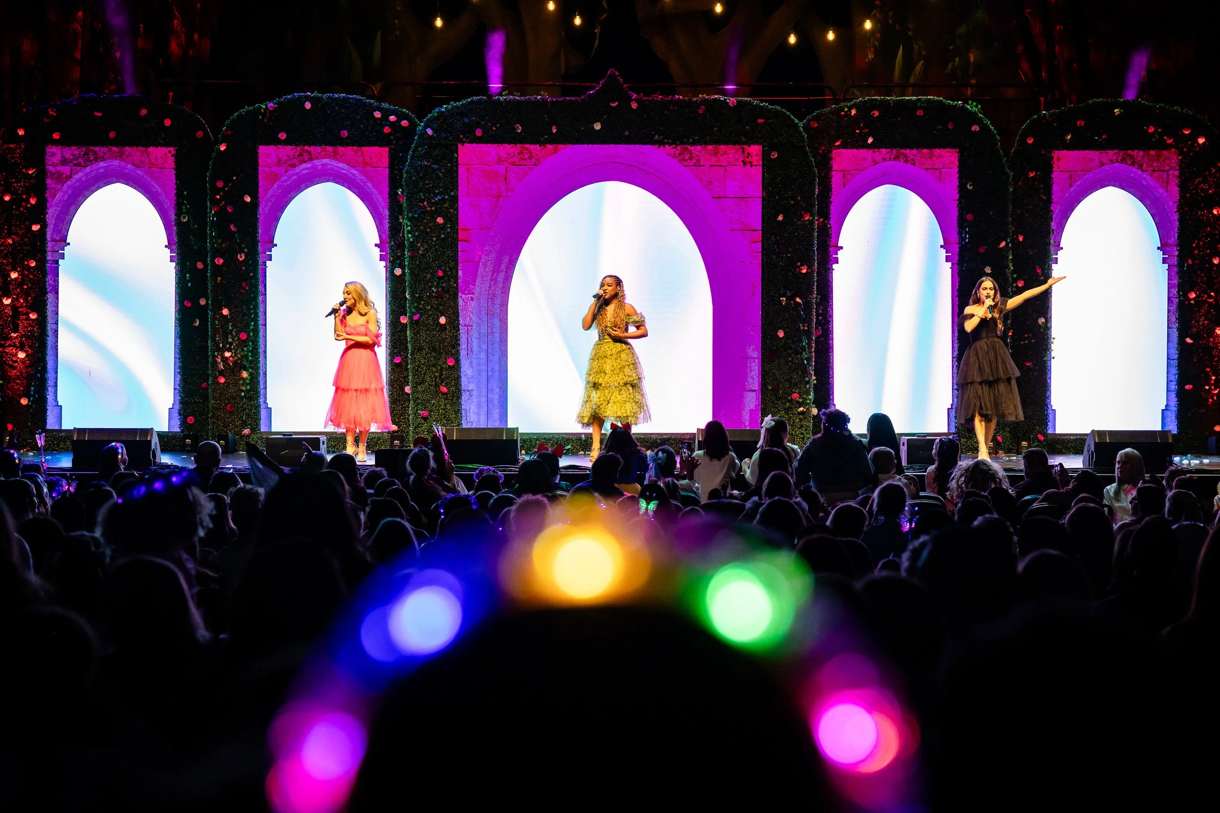 Three women on stage in colorful dresses singing into microphones, with a decorated background of arches and lights, audience in front with colorful bokeh effect.