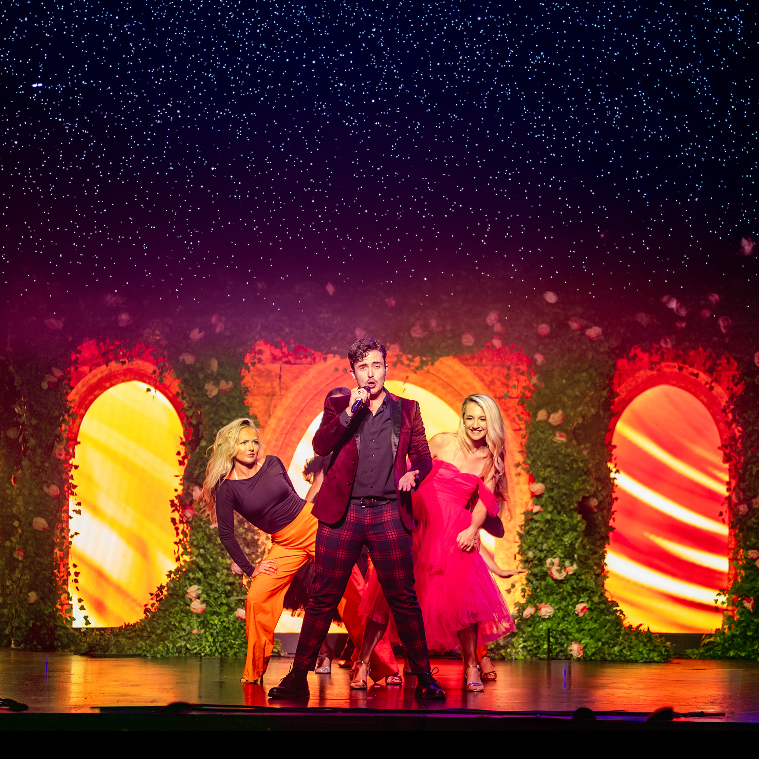A group of performers standing on a stage with a colorful, floral, and illuminated backdrop under a starry sky. There are mostly women and one man, with the man at the center holding a microphone. They appear joyful and engaged in a performance.