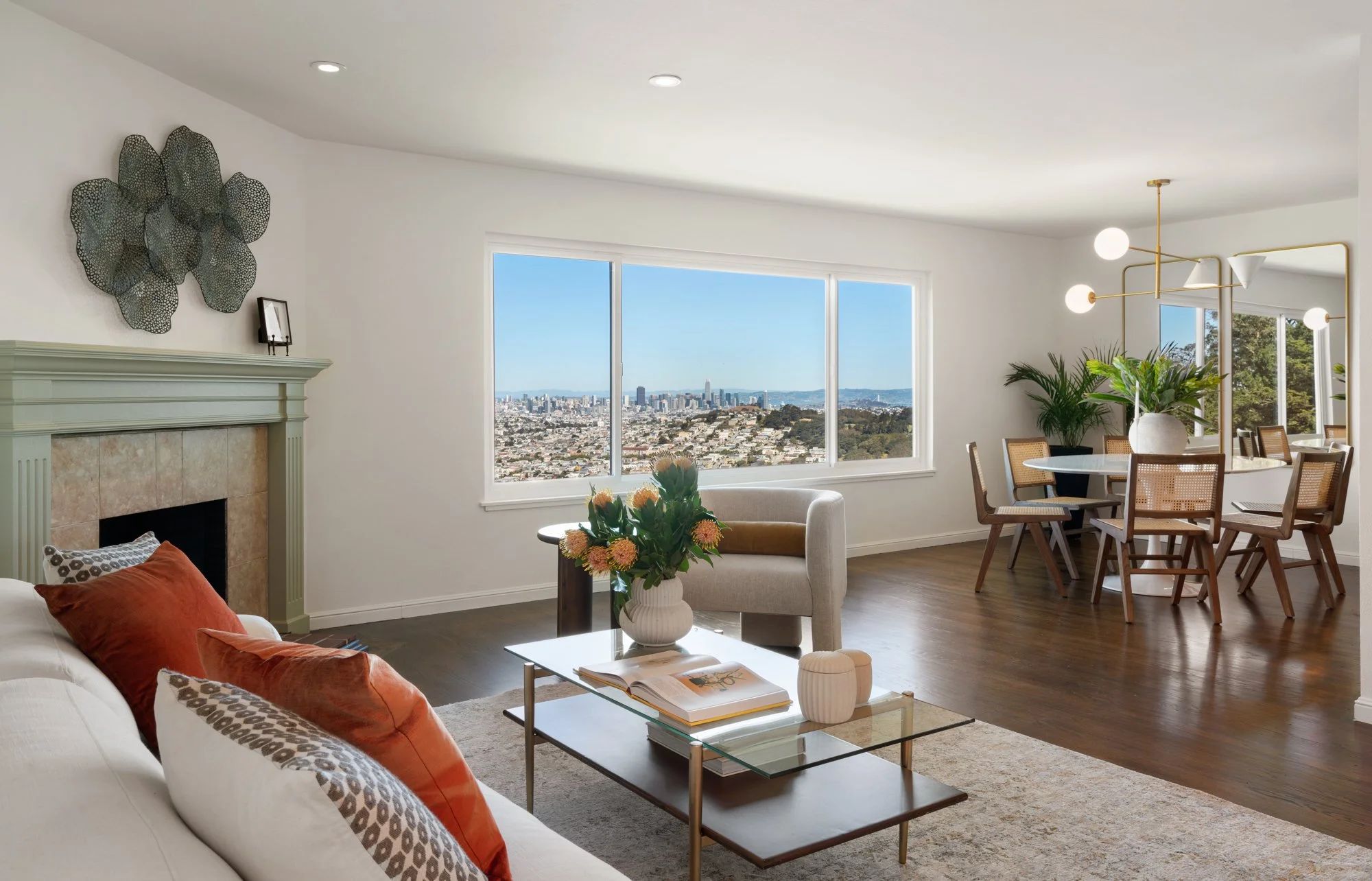 Living room with large window view of city skyline, white sofa with orange and patterned pillows, glass coffee table with books and decorative items, fireplace with green mantel, dining area with round table and chairs, modern chandelier, and indoor plants.