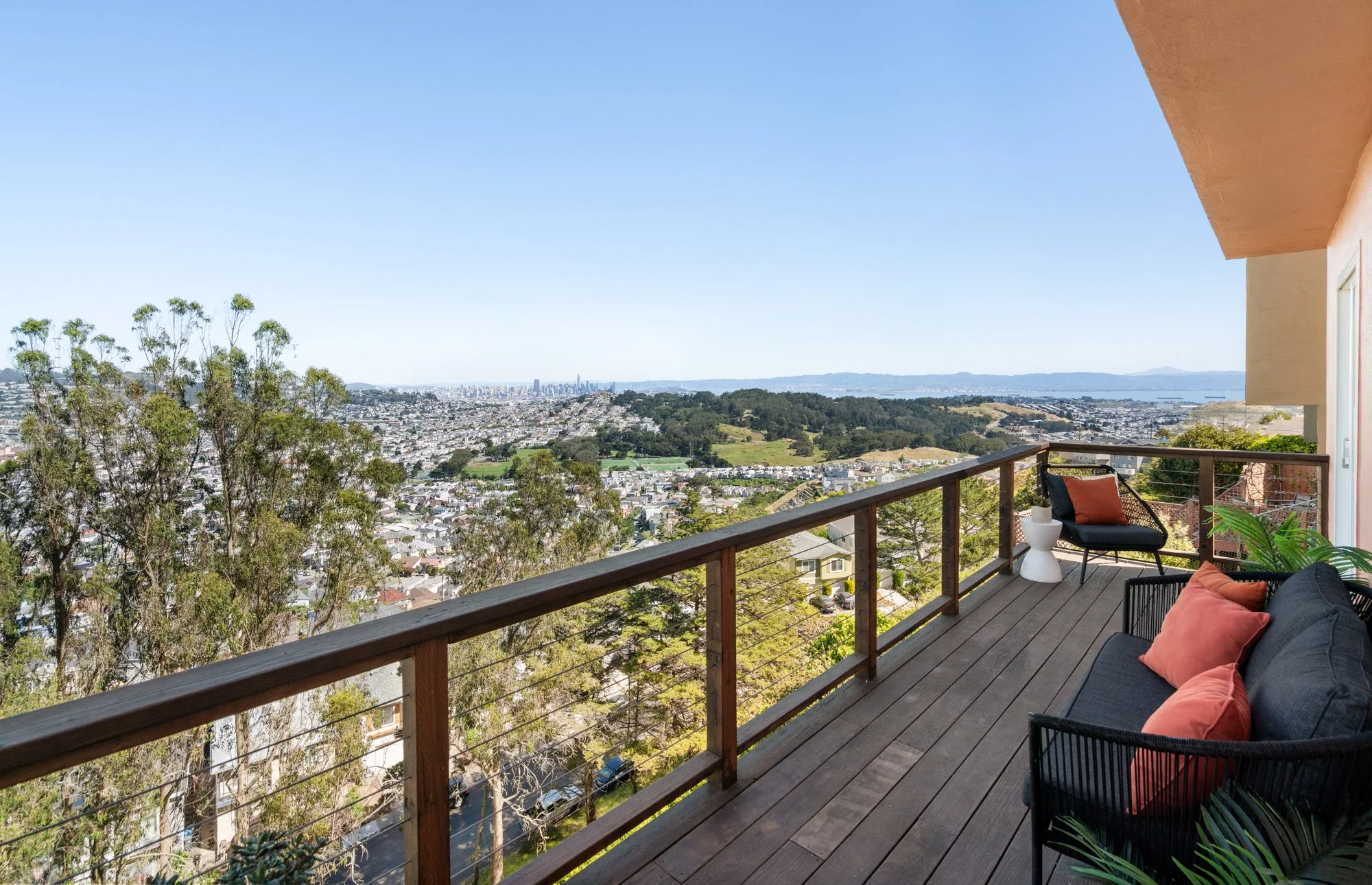 Balcony overlooking a city and hilly landscape under a clear blue sky, furnished with black outdoor seating, colorful cushions, and a small white side table.
