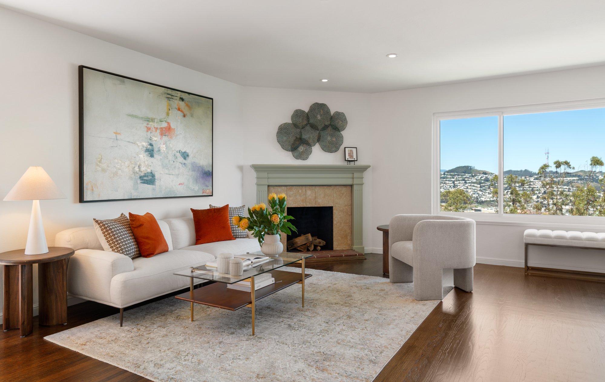Living room with white couch, orange and patterned pillows, glass coffee table, outdoor view through large window, fireplace, abstract wall art, beige armchair, side table, and decorative wall art.