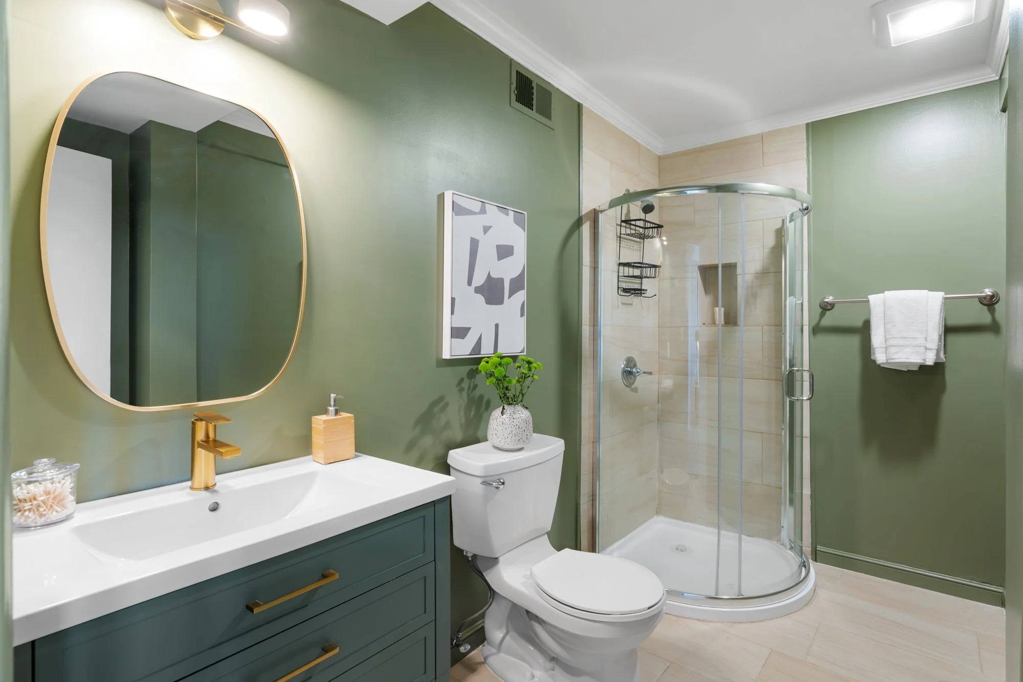 Modern bathroom with green walls, gold accents, a white sink with green cabinetry, a mirror, a toilet with a plant on top, a glass shower enclosure, and a towel on a bar.