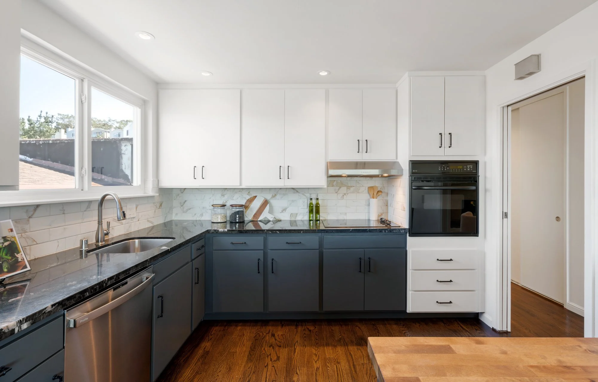 Modern kitchen with white upper cabinets, dark lower cabinets, marble backsplash, black countertop, hardwood floor, and a window above the sink.