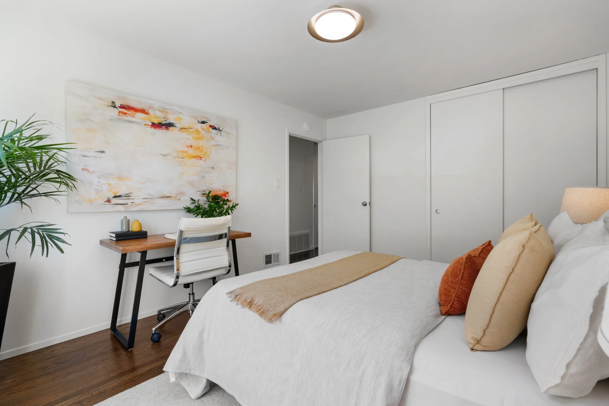 A bedroom with a white bed, beige and rust-colored pillows, a beige throw blanket, a wooden desk with a white chair, a potted plant, and an abstract painting on the wall.