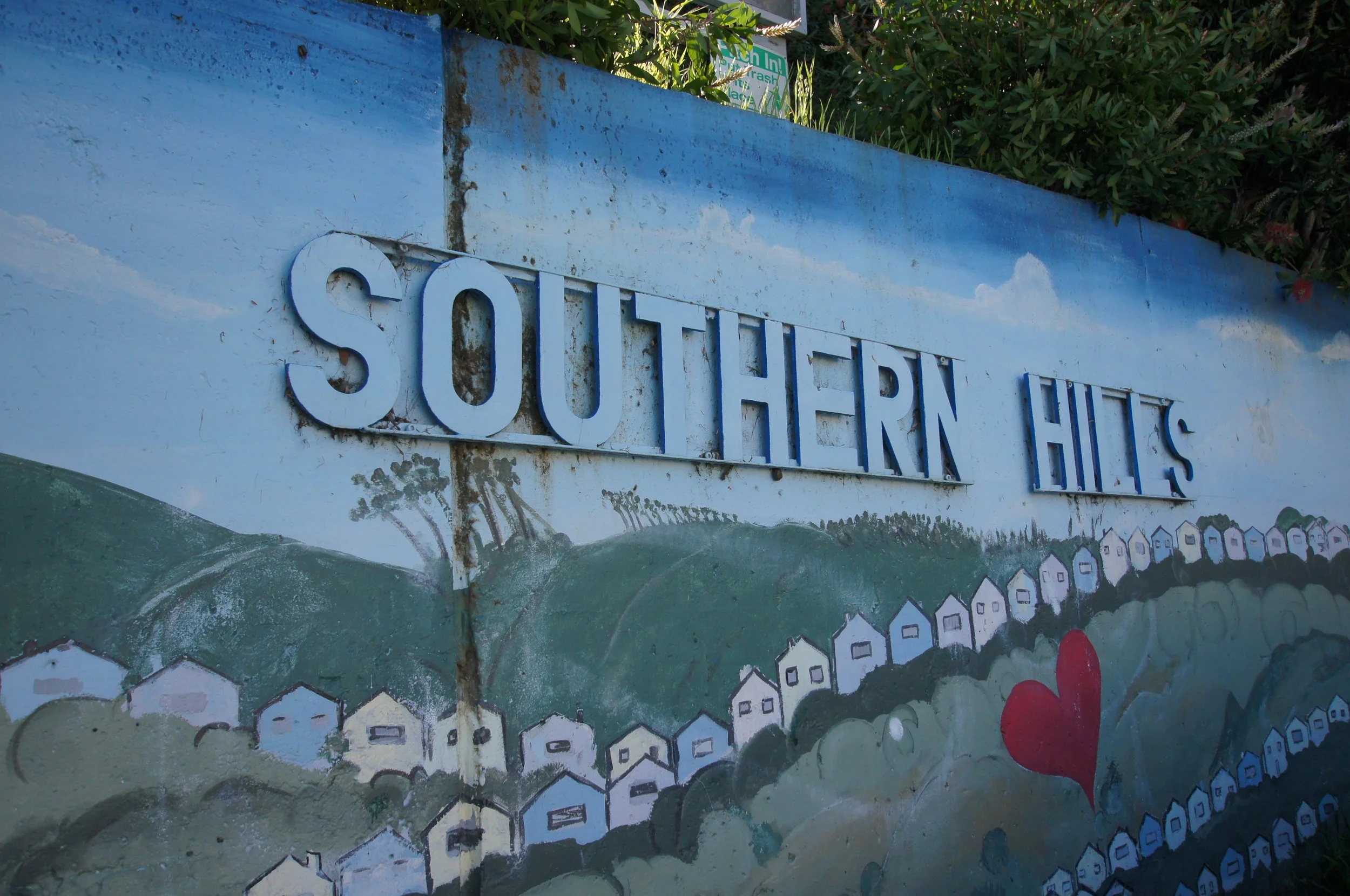 A mural with the words 'Southern Hills' written in large blue letters on a painted background of green hills and a blue sky with clouds. There are small houses painted at the bottom of the mural and a red heart near the houses.