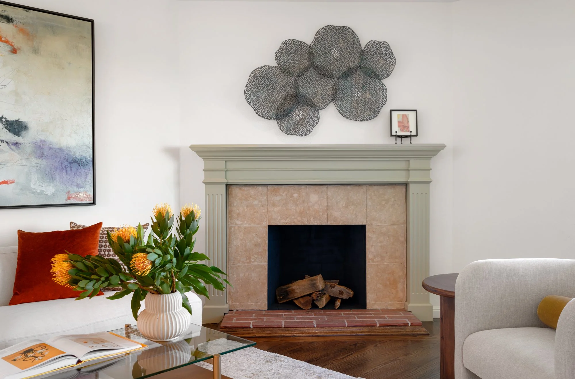 Living room with a white sofa, a glass coffee table, a vase with yellow flowers, a fireplace with beige tiles and an abstract wall art above it, and a small framed picture on the mantelpiece.