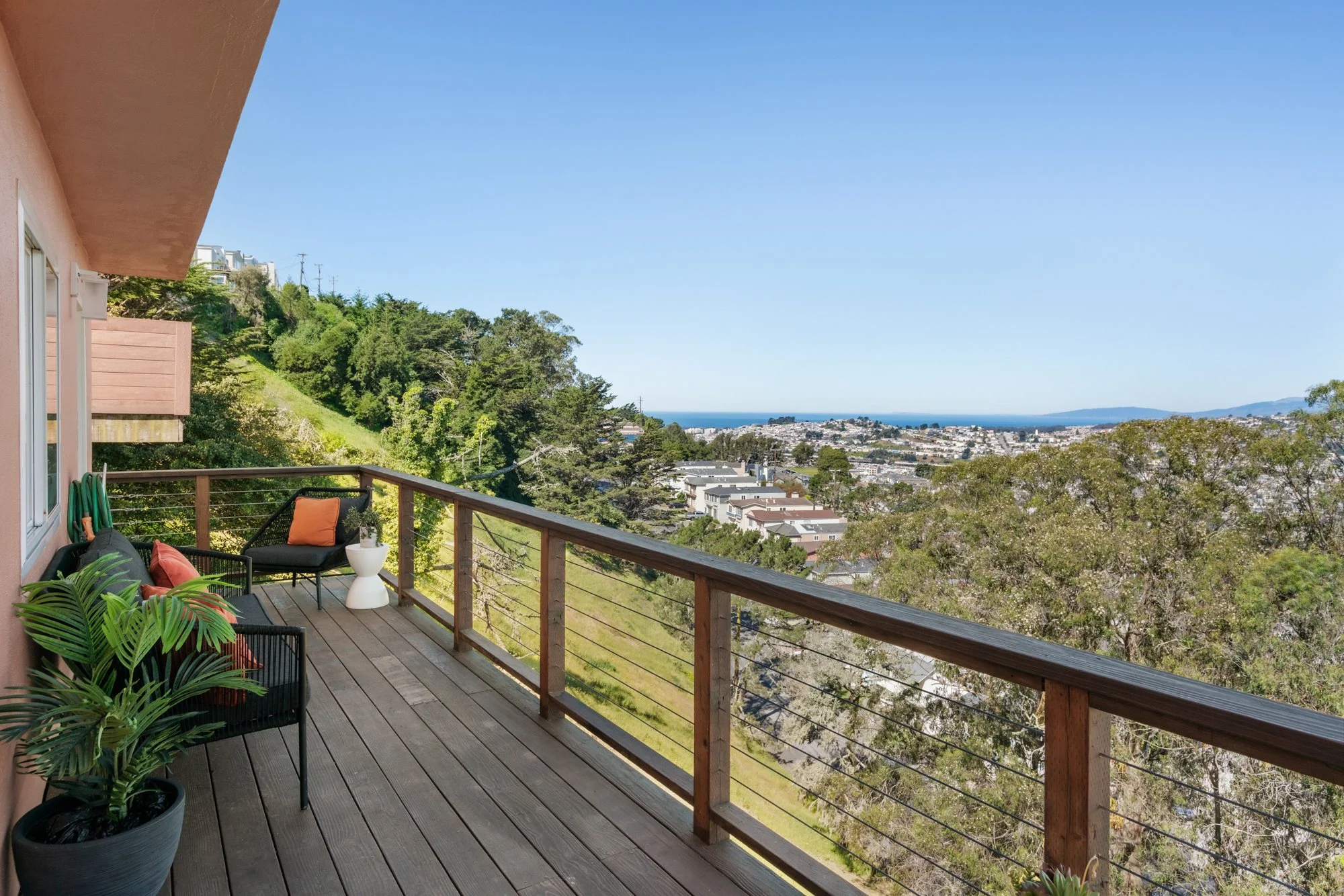 Balcony with outdoor seating overlooking a hillside and distant cityscape, with blue sky and ocean in the background.