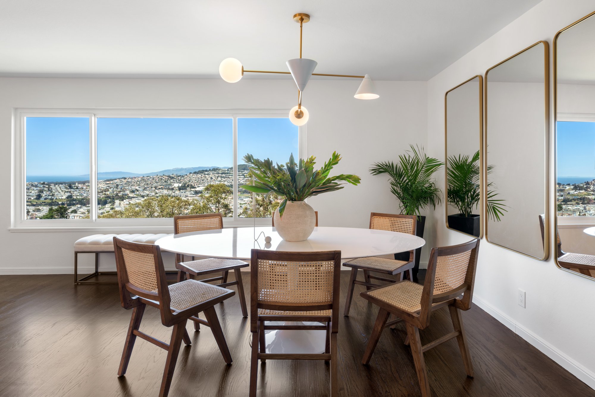 Modern dining room with large window view, round white table, six wooden chairs with cane seats, decorative plants, and gold-framed mirrors.