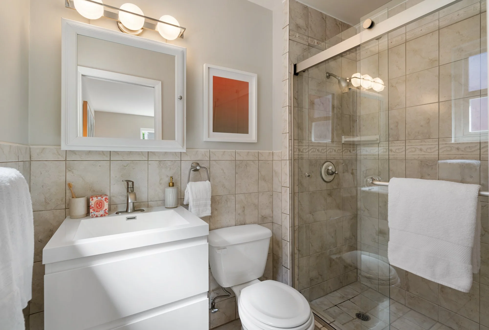 Bathroom with white vanity, mirror, towel, soap dispenser, toothbrush holder, toilet, and glass-enclosed shower with beige tiles and towel rack.