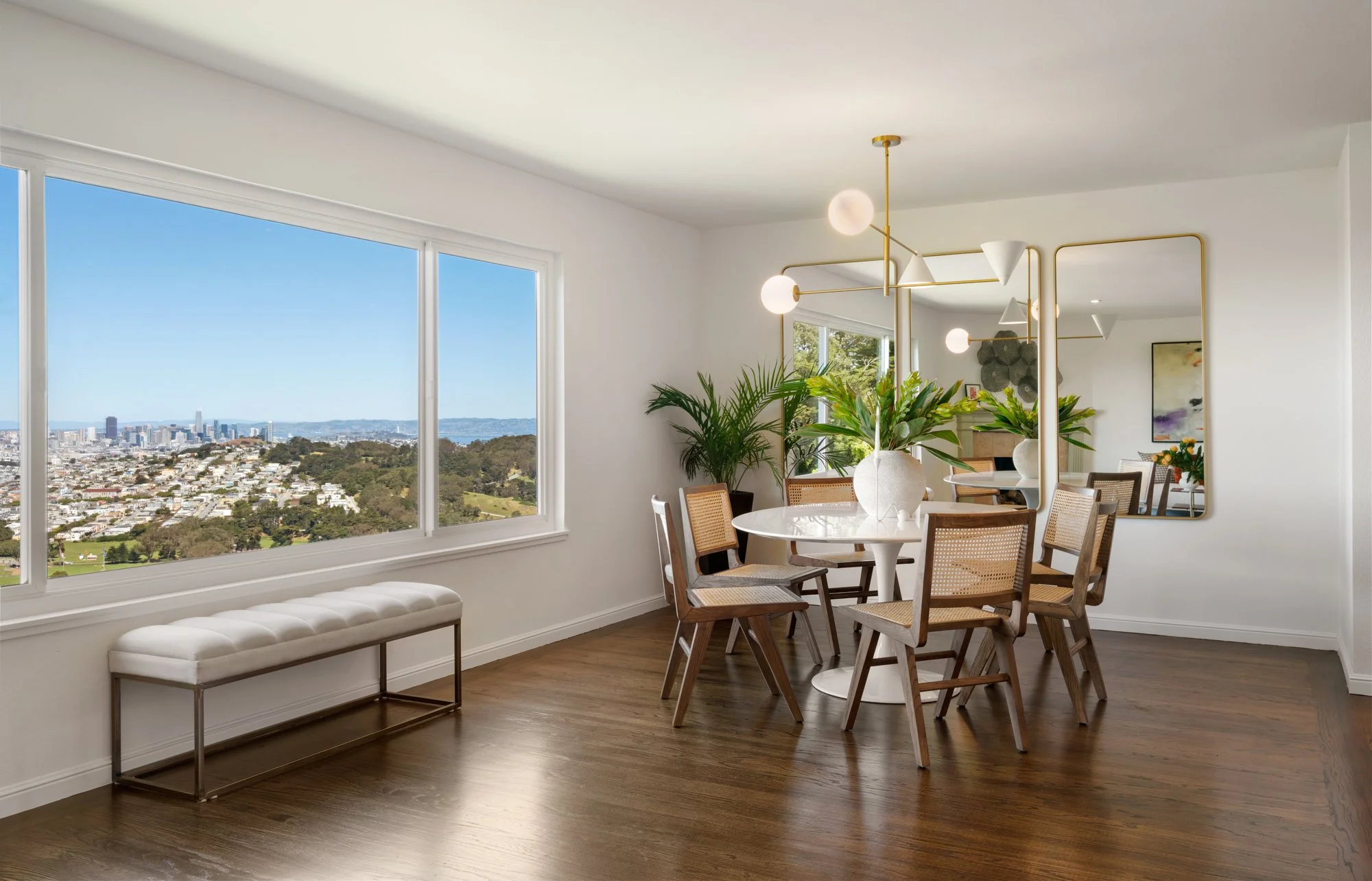 Bright dining room with large window showing city view, wooden floor, round table with white base, six wooden chairs with cane backs, large green plants, modern chandelier, and a large mirror on the wall.