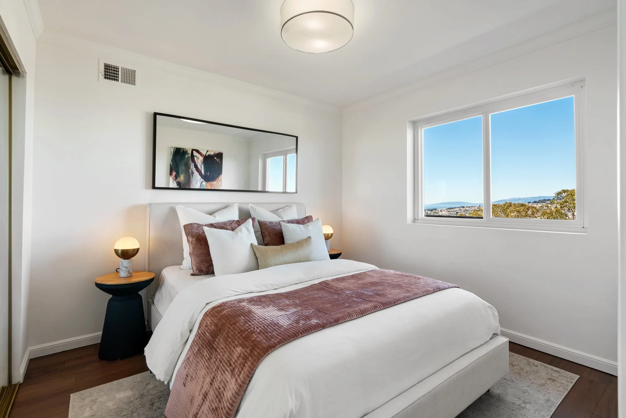 Bright modern bedroom with a large bed, pink and white bedding, wooden nightstands with lamps, a large mirror, and a window showing a scenic view of trees and mountains.