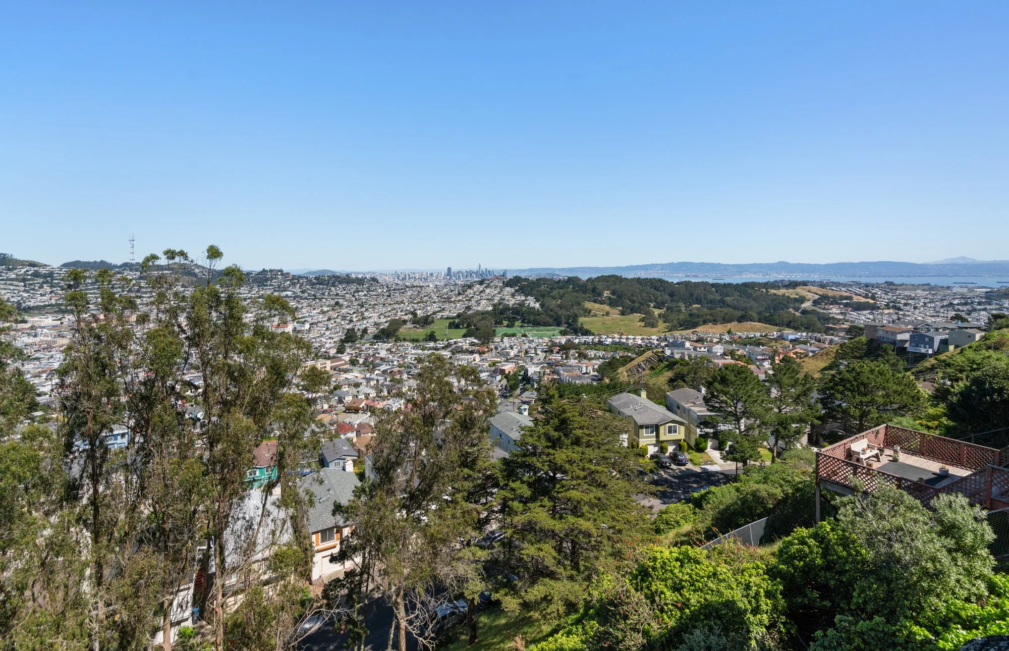 A scenic view of a cityscape from an elevated vantage point, showing a mix of residential houses, trees, and rolling hills under a clear blue sky.