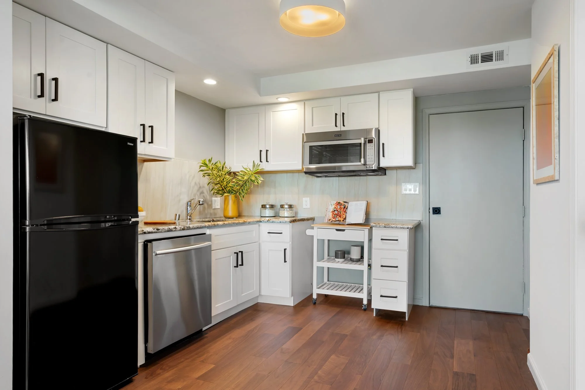 Modern kitchen with white cabinets, stainless steel appliances, a black refrigerator, microwave, and a small white rolling cart. Decor includes a yellow vase with green plants and candles on the counter.