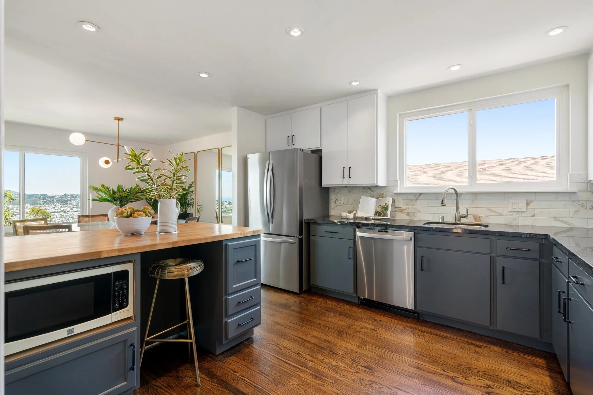 Modern kitchen with white upper cabinets, dark lower cabinets, stainless steel appliances, wooden countertop island with a plant, open layout, hardwood floors, and large window letting in natural light.