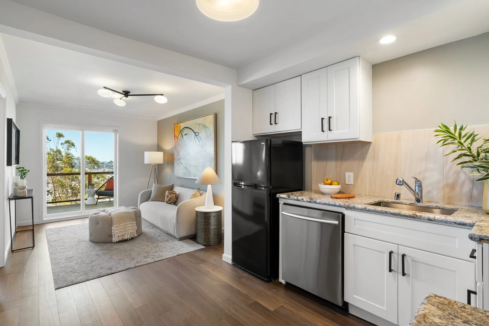 Modern living room with a sofa, artwork, and balcony view, connected to a kitchen with white cabinets and granite countertops.