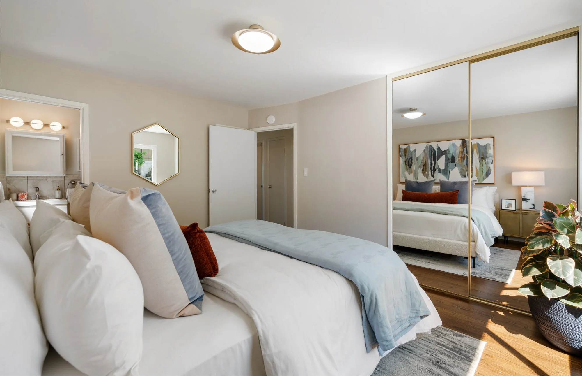 A modern bedroom with a bed, pillows, a closet with mirrored sliding doors, and a nightstand with a lamp and decor, reflected in the mirrored closet.