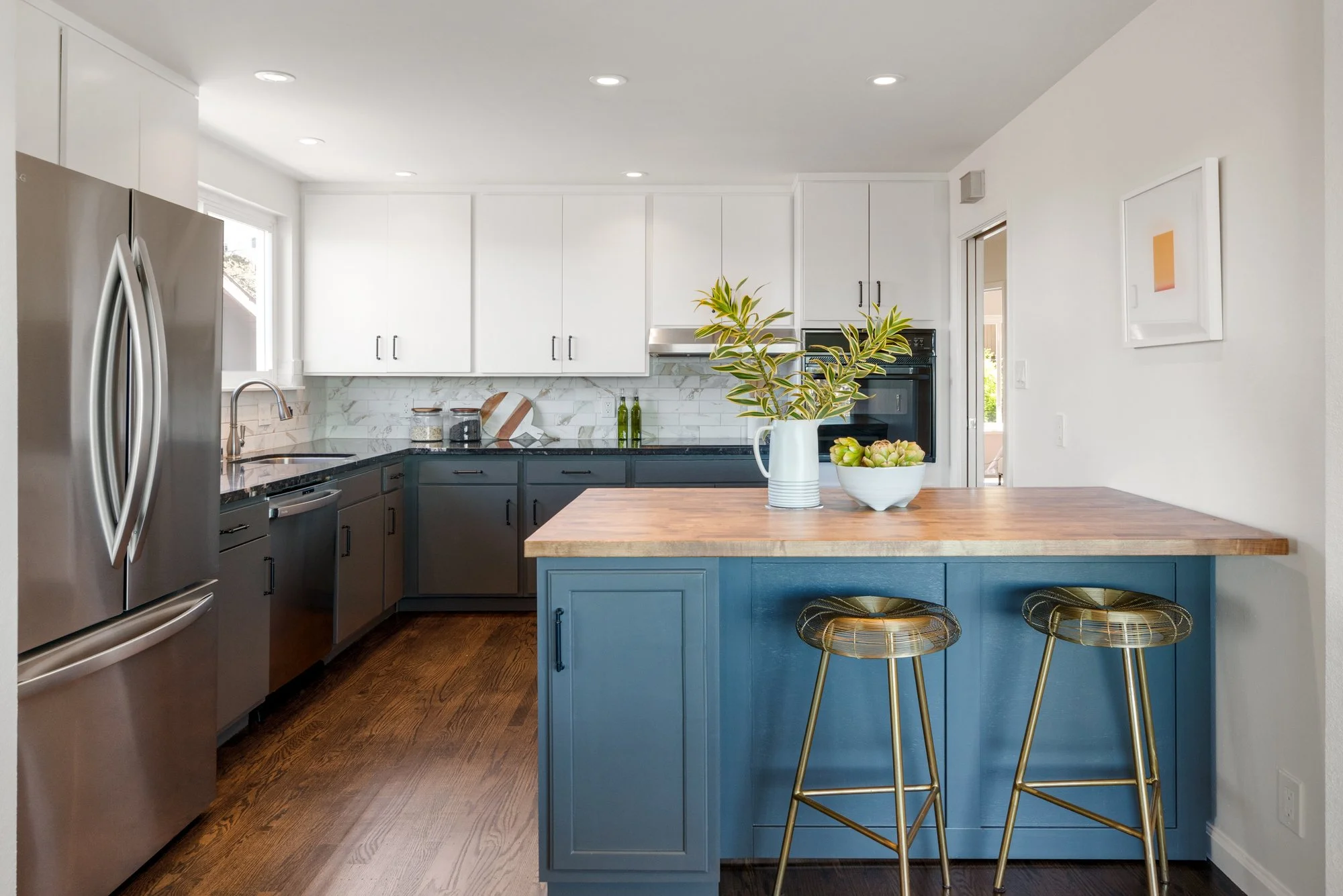 Modern kitchen with stainless steel refrigerator, white and dark gray cabinets, wooden countertop on a blue kitchen island, and two gold bar stools.