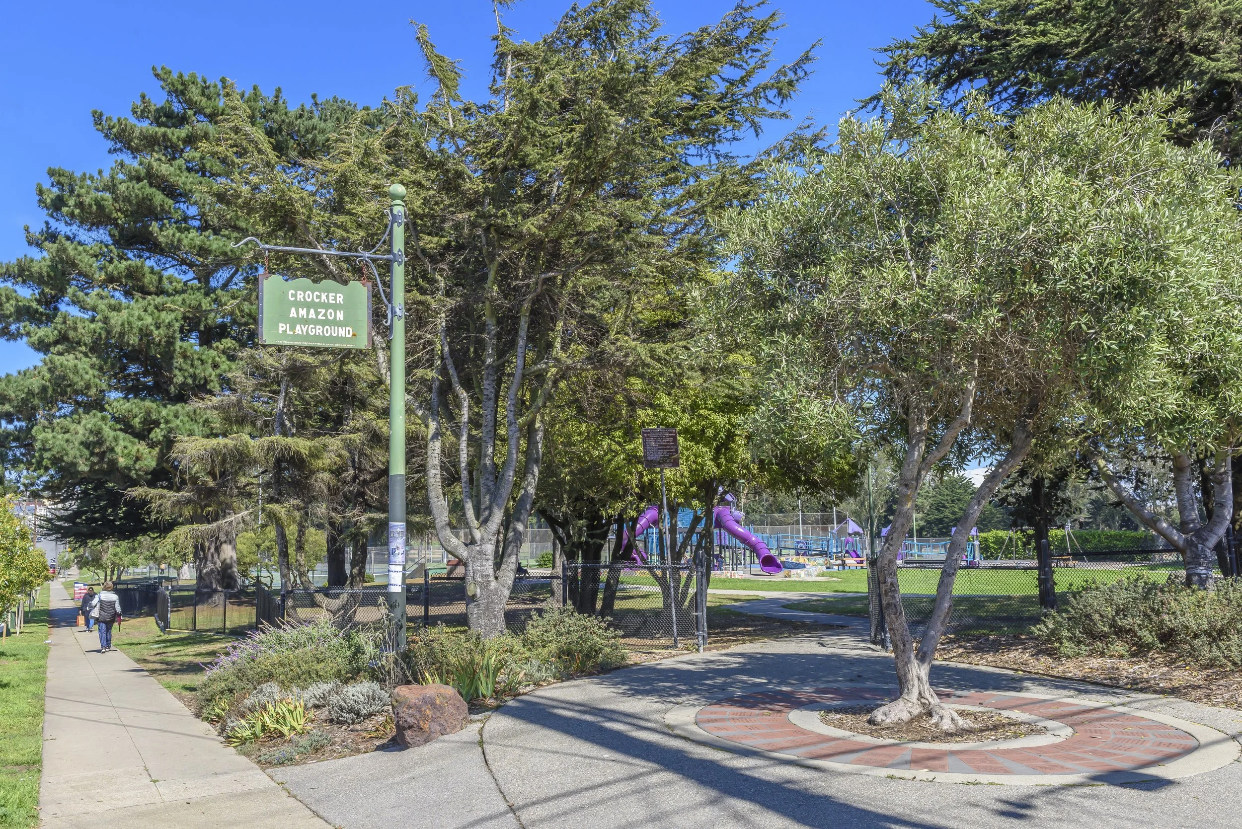Crocker Amazon Playground with trees, a chainlink fence, a paved pathway, and purple slides in the background.