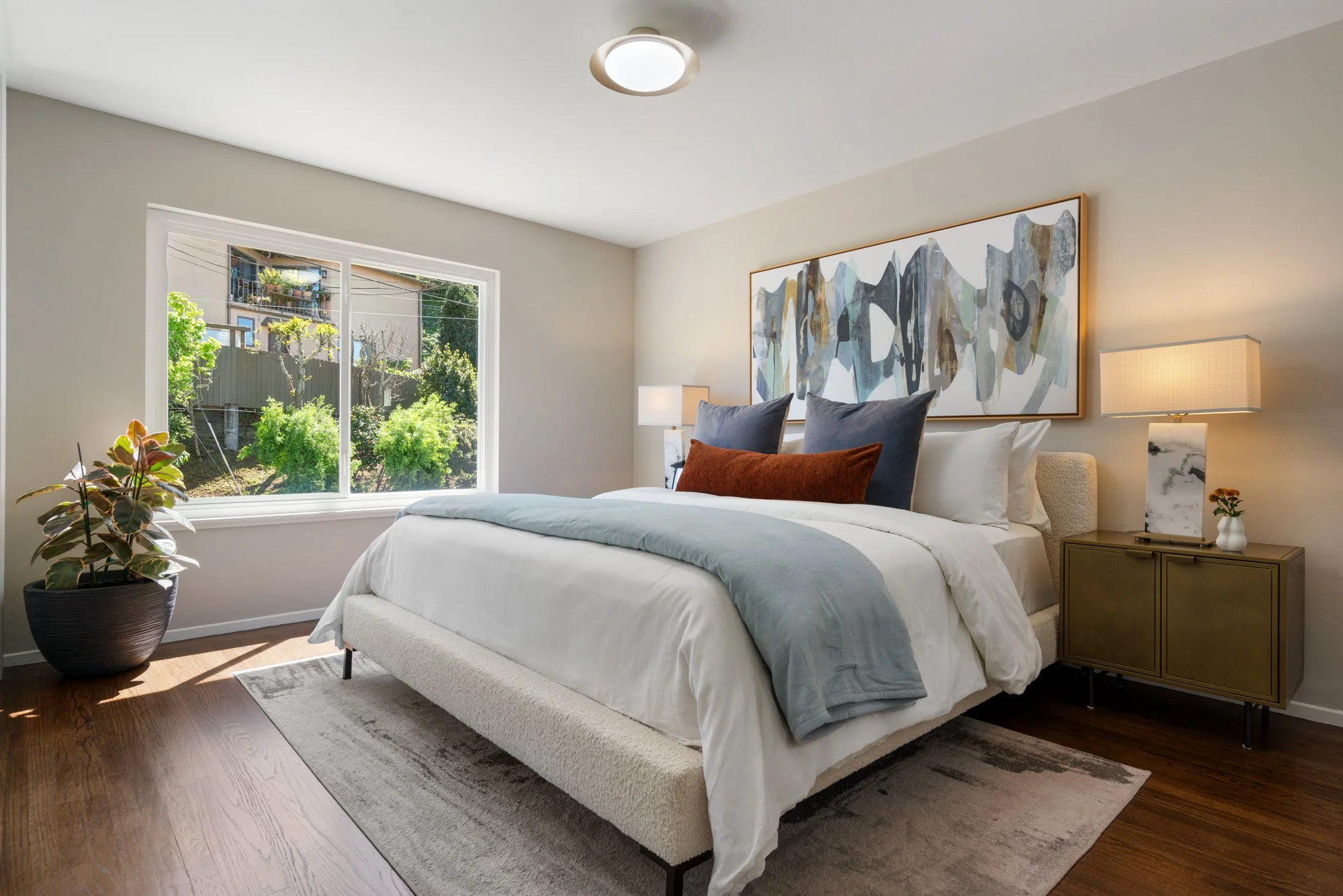 A bedroom with a large bed, white bedding, and decorative pillows. Window showing greenery outside, a modern dresser with a lamp, a framed art piece above the bed, and a potted plant in the corner.