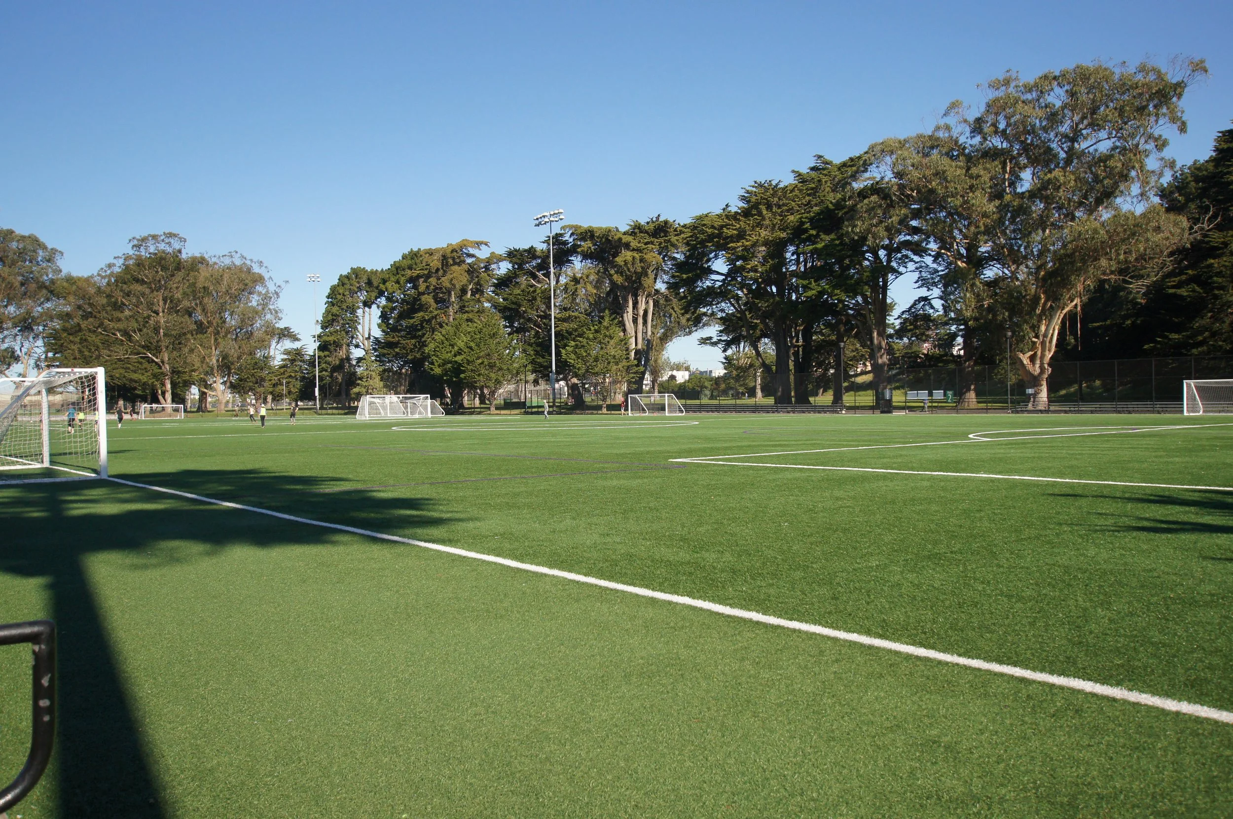 A soccer field with goalposts, surrounded by trees under a clear blue sky, with players practicing in the distance.