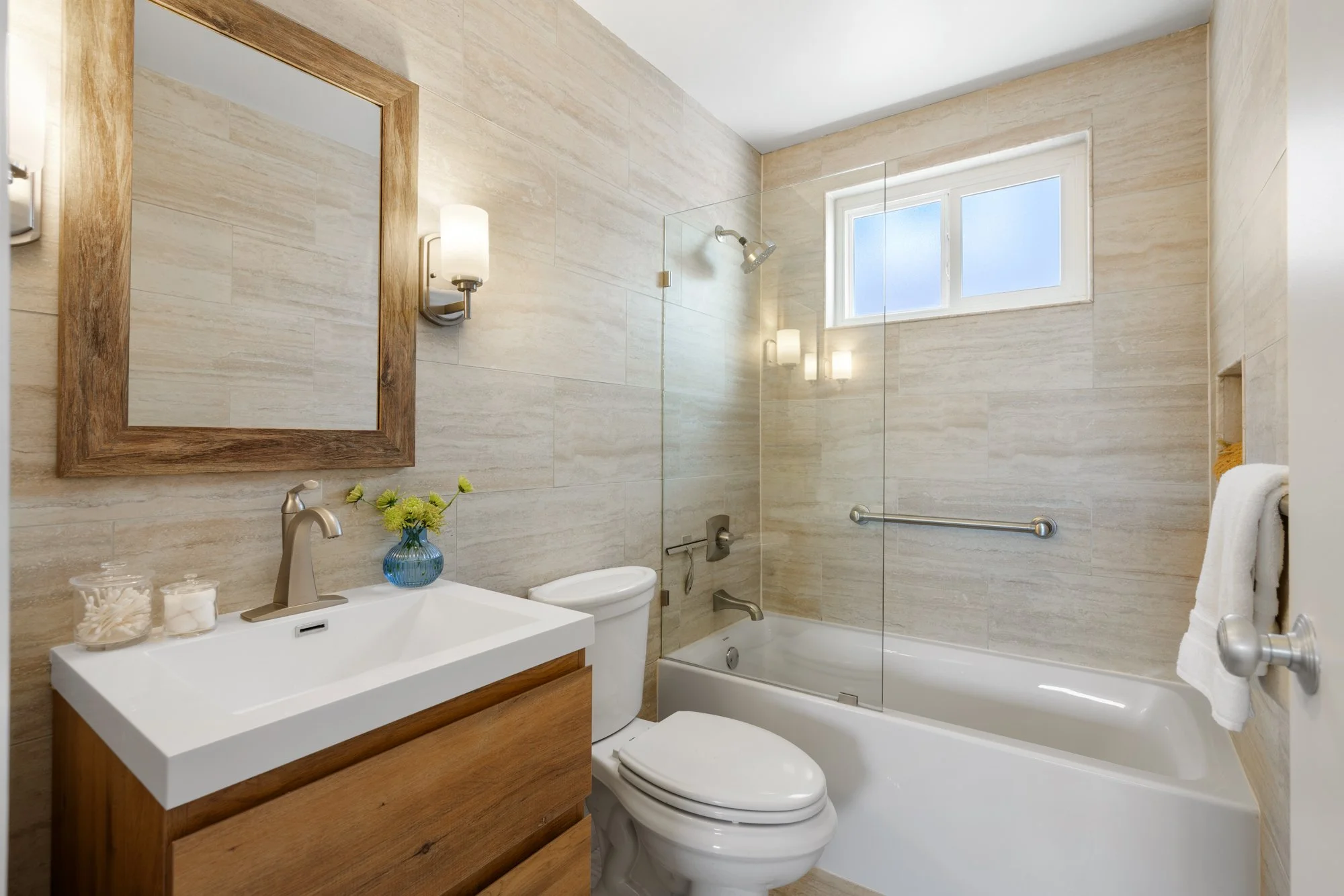 Bathroom with beige tiled walls, a white bathtub with a glass partition, a window above the tub, a toilet, and a wooden vanity with a white sink and a mirror.