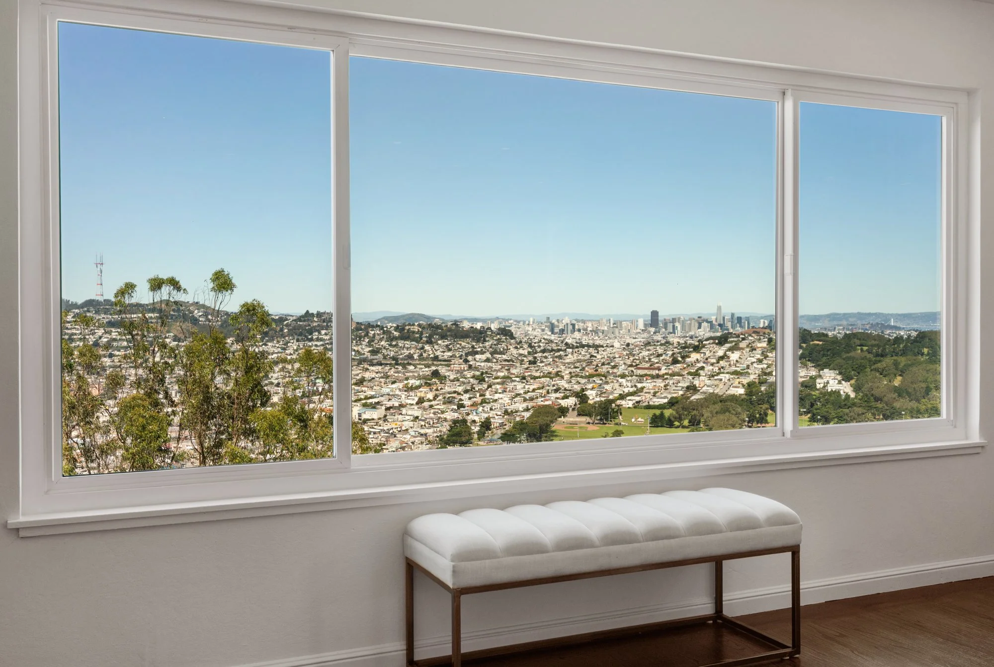 View of a cityscape through a large window with three panes, from inside a room with a white bench by the window.