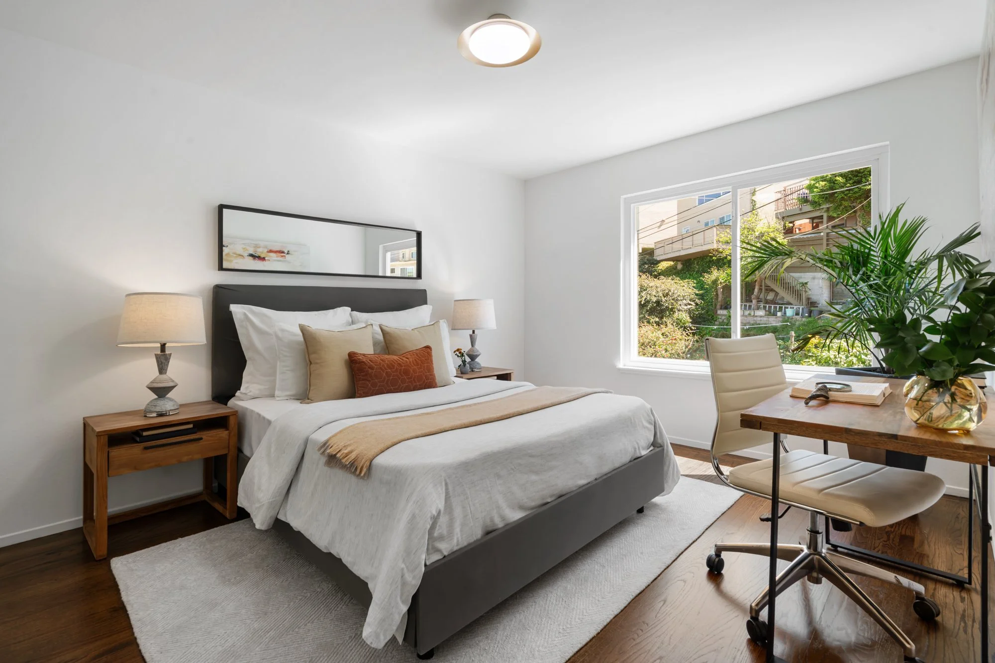 Modern bedroom with a bed, two nightstands with lamps, a large mirror, a window showing greenery, a desk with a chair, a potted plant, and hardwood floors.