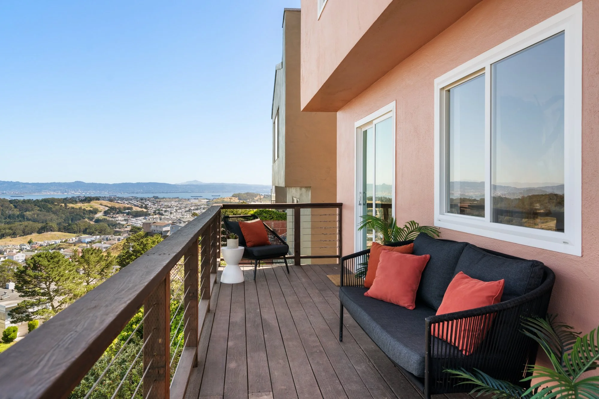 Balcony with black outdoor furniture, potted plants, and scenic view of hills, water, and houses in the distance.