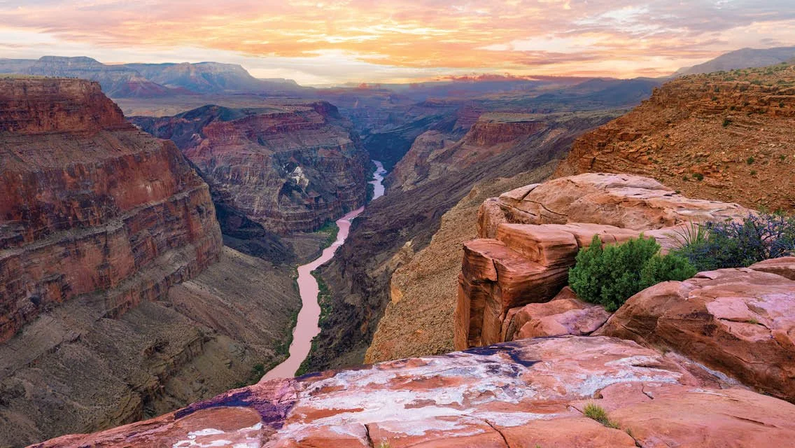 Sunset over the Grand Canyon with river flowing through the canyon, rugged rocks in foreground and layered cliffs in the background.