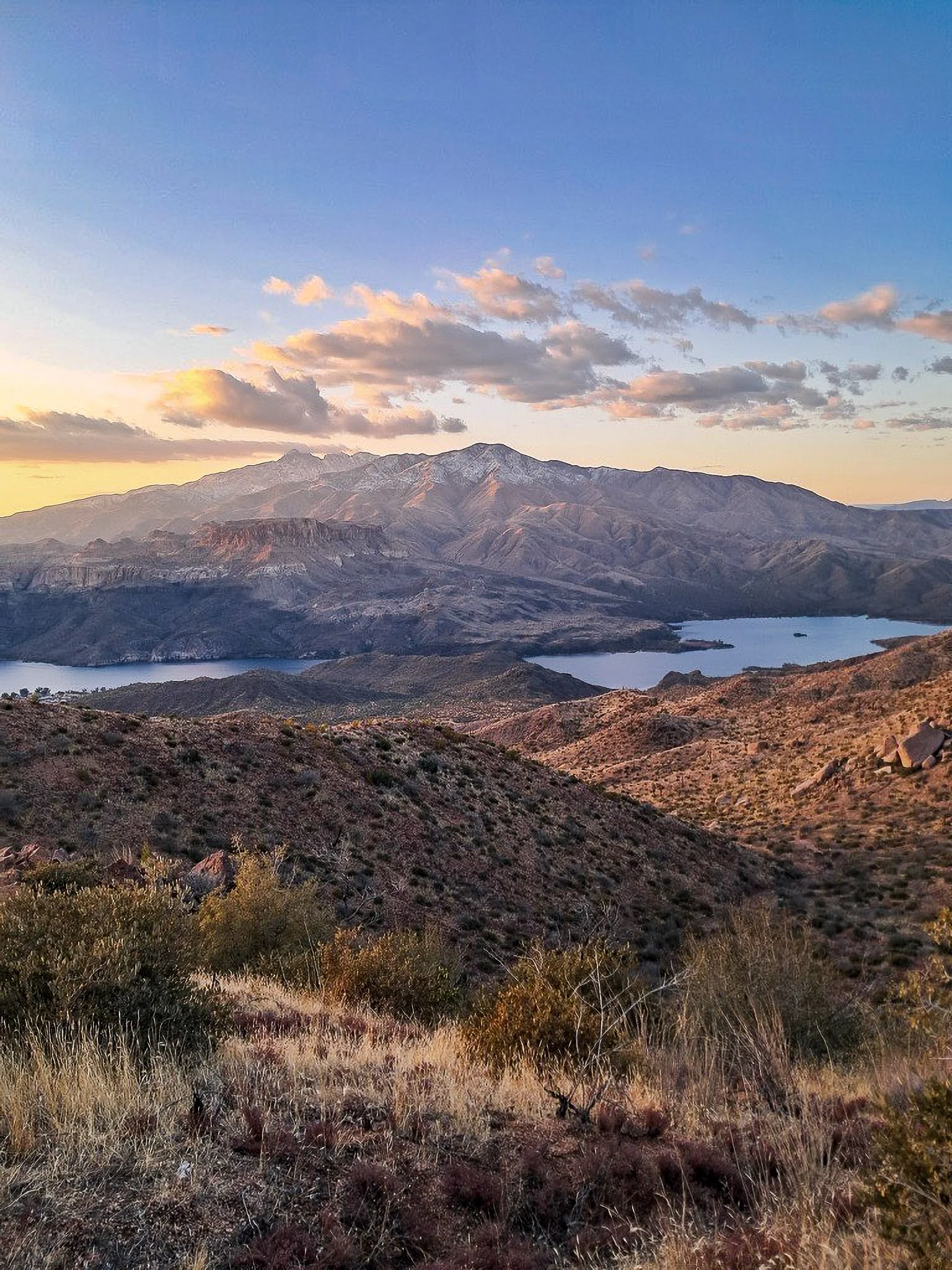 A landscape view of a mountainous area during sunset with a lake, rugged terrain, and dry, brownish vegetation in the foreground.