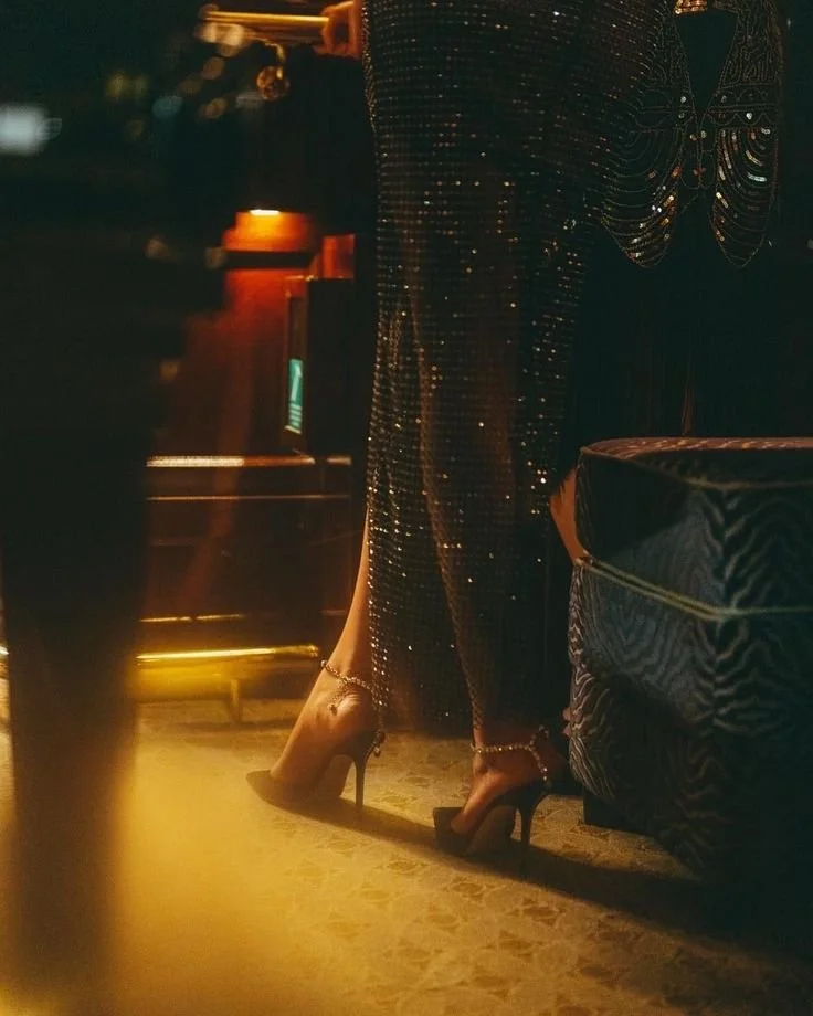 Woman wearing sparkling, sequined dress and high heels standing near a velvet armchair in a dimly lit room.