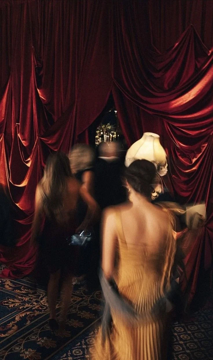 People entering a room with red velvet curtains, dim lighting, and a vintage lamp, at what appears to be an upscale event or party.