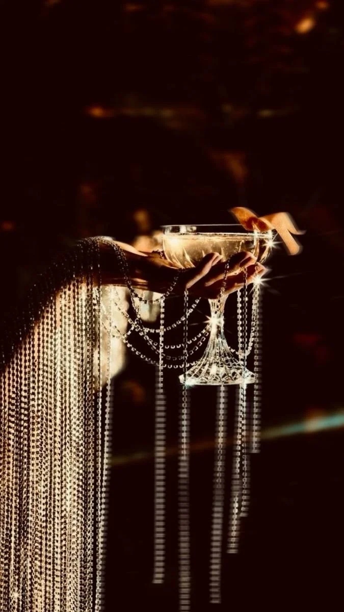 A hand holding a cocktail glass with a decorative element, surrounded by shimmering beads or jewelry, against a dark background with warm lighting.