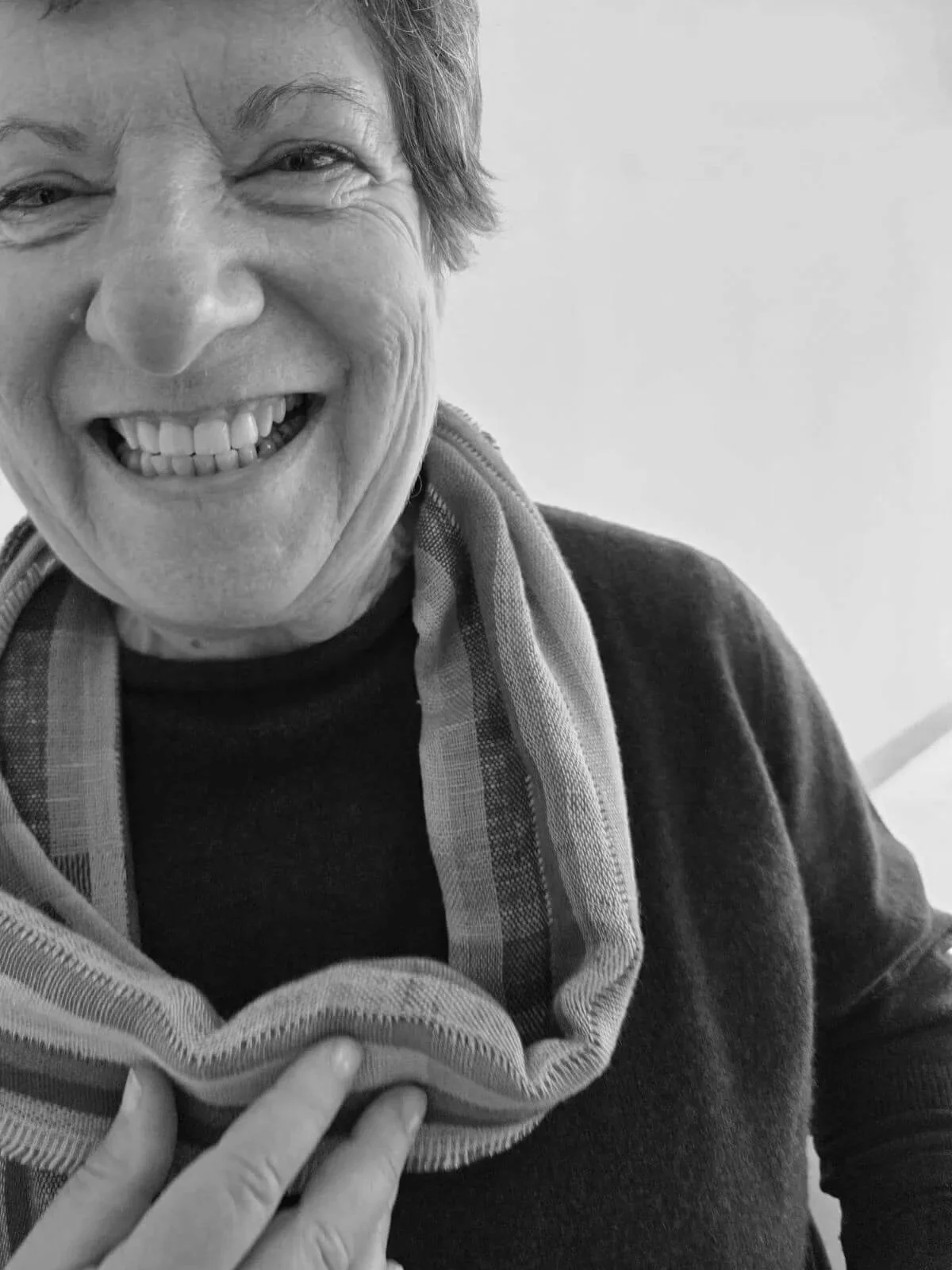 Close-up black and white photo of smiling elderly woman with short hair, wearing a dark top and a patterned scarf.