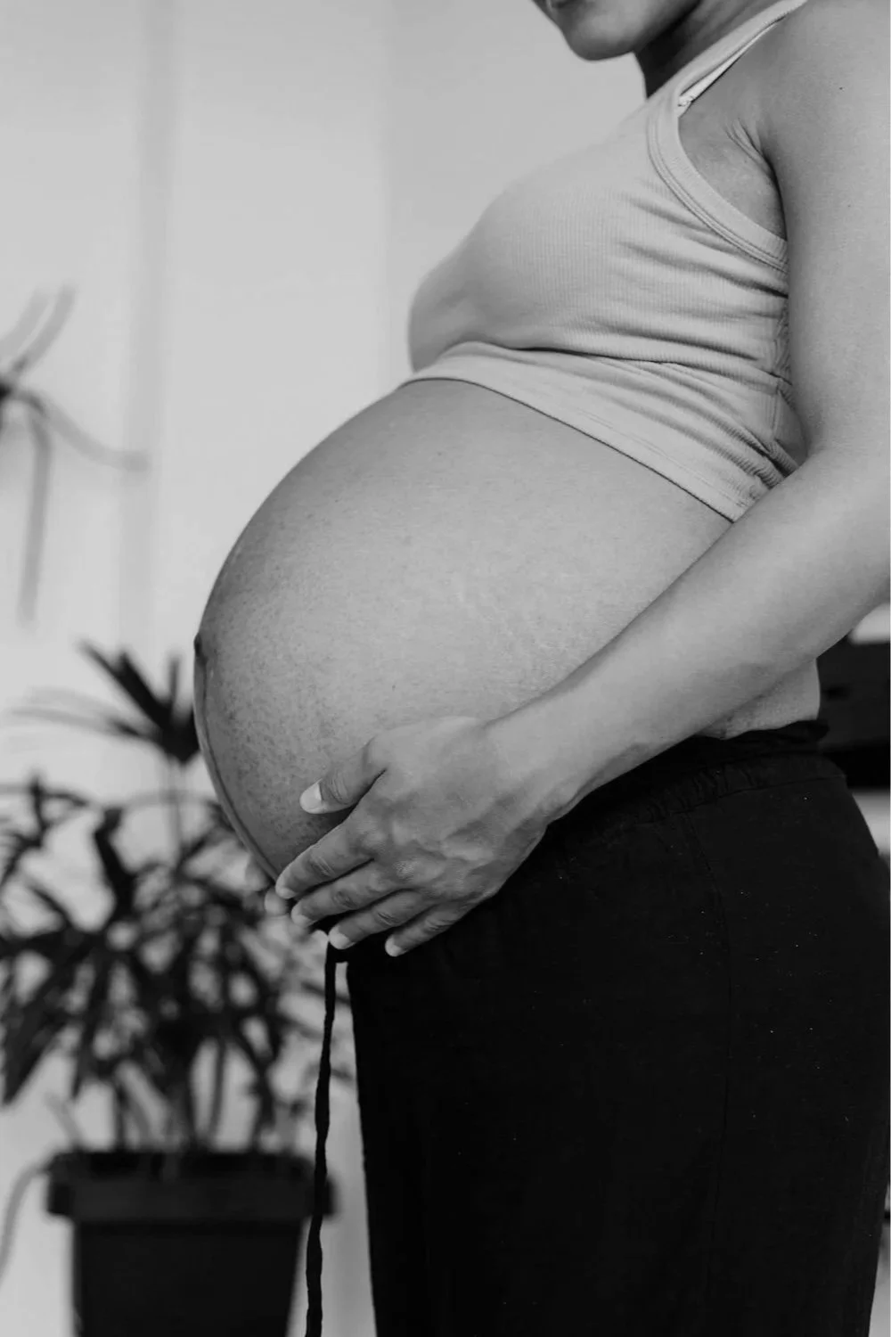 Close-up of pregnant woman holding her belly with both hands, wearing a tank top and pants, in black and white.
