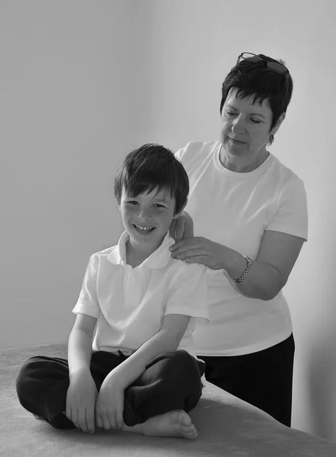 A woman giving a neck massage to a young boy sitting cross-legged on a massage table in a room with plain walls.