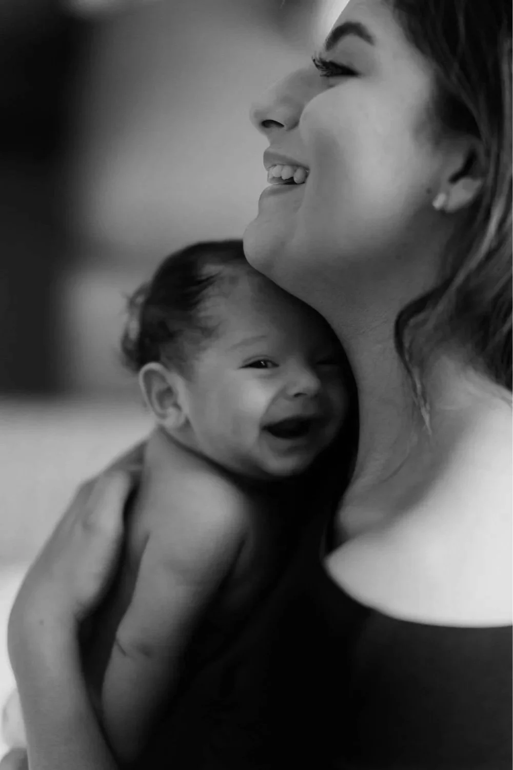 A woman smiling in close-up with a baby resting on her shoulder, both sharing joyful expressions. The image is in black and white.